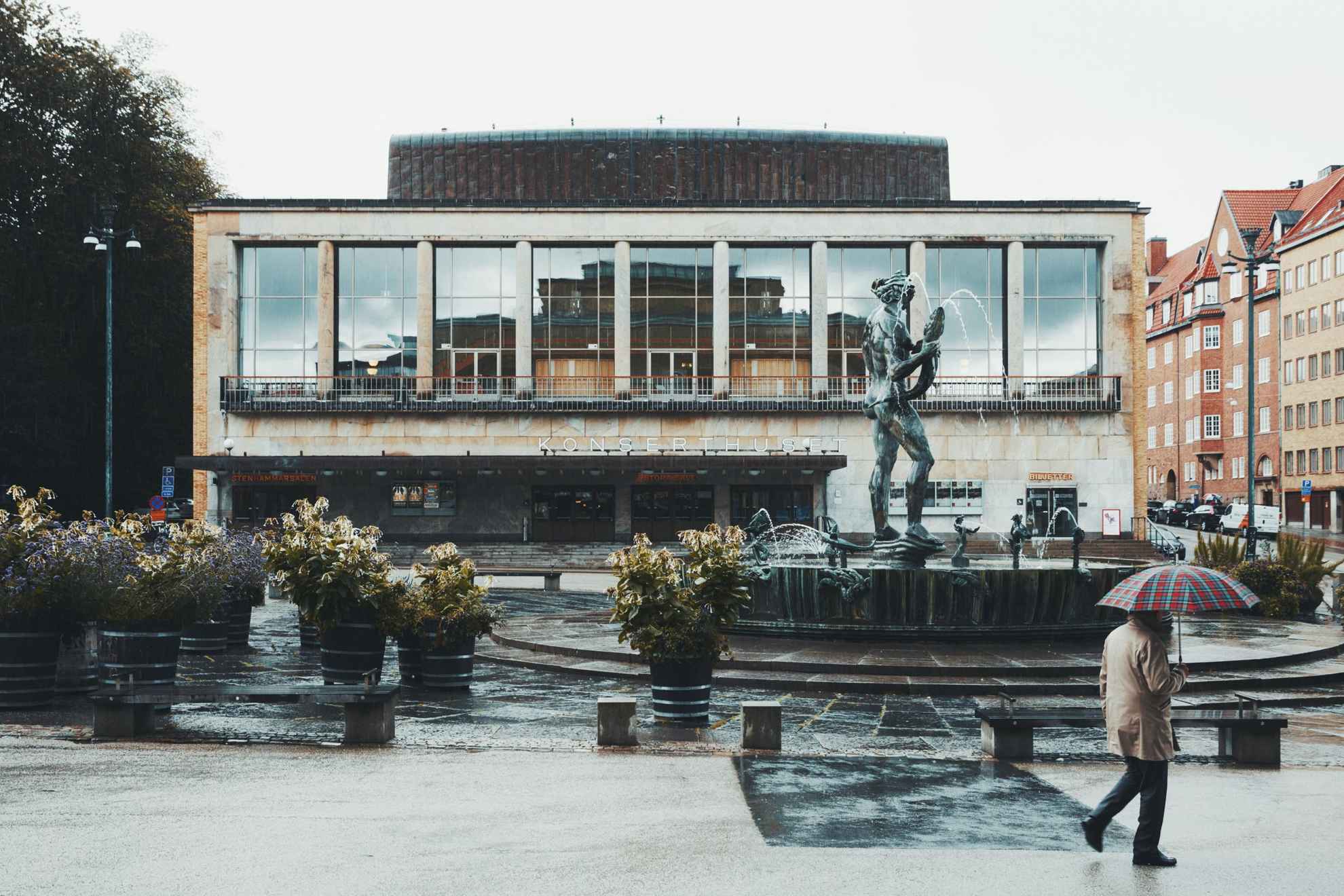 A man holding an umbrella walks past the Concert hall and Poseidon statue in Gothenburg.