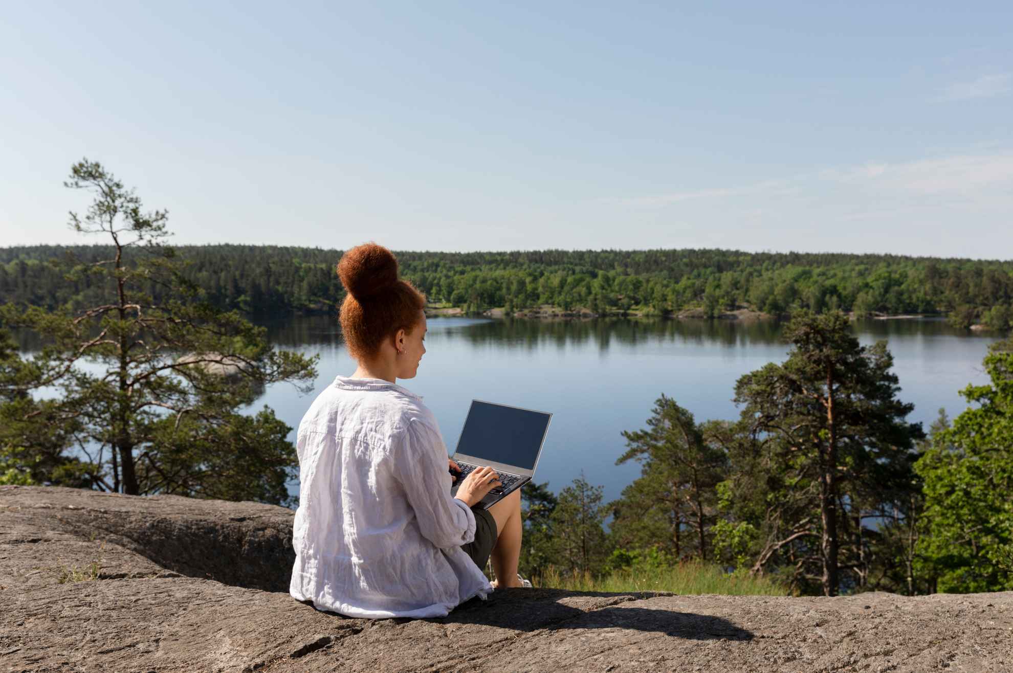 Eine Person sitzt mit ihrem Laptop auf einem Felsen vor einem Waldsee
