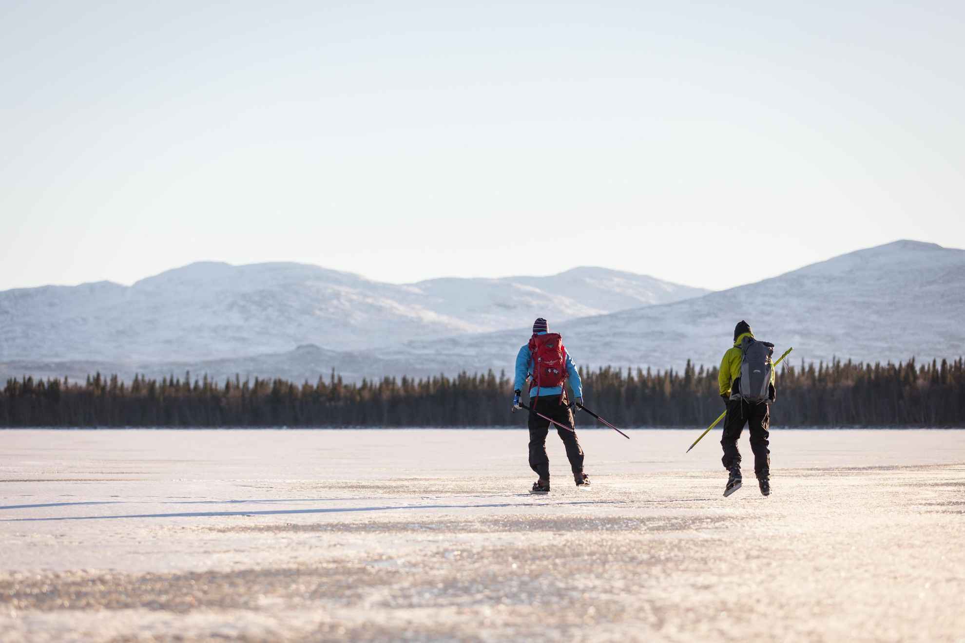 Zwei Männer laufen Schlittschuh auf einen zugefrorenen See vom Fotografen weg. Im Hintergrund eine Bergkette.