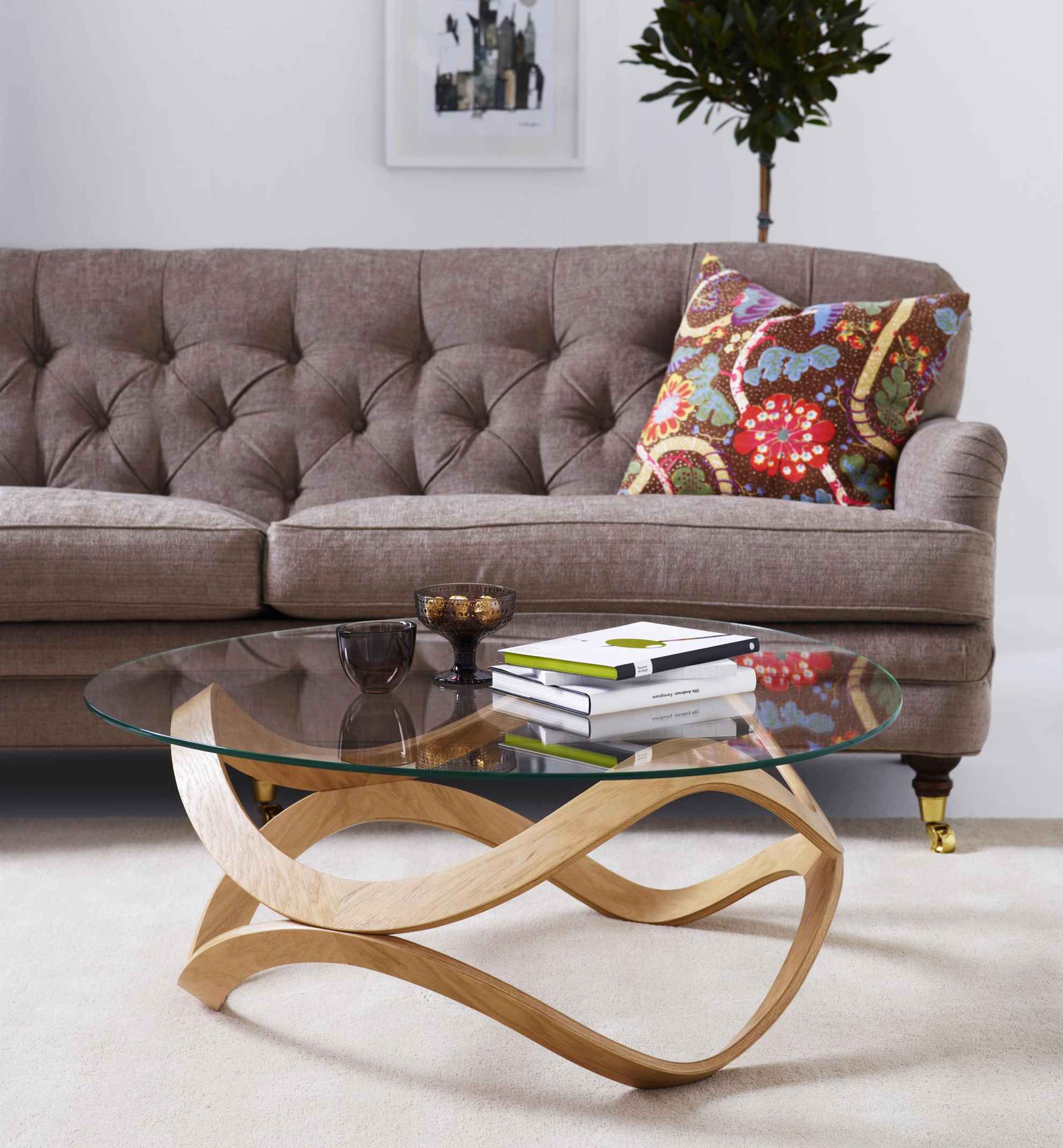 Ein rund gestalteter Glastisch vor einem braunen Sofa. Auf dem Sofa liegt ein gemustertes Kissen.