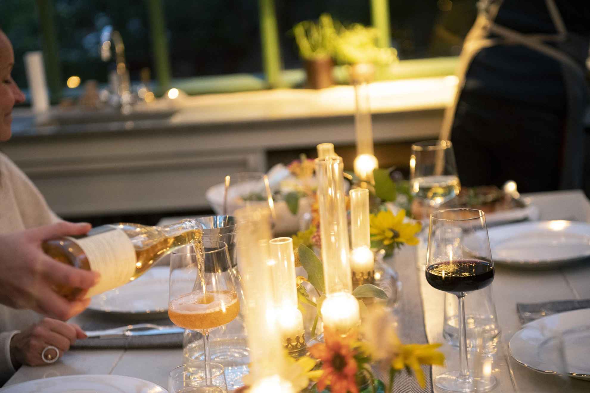 Ein Esstisch mit Gläsern, Besteck, Tellern, Blumen und brennenden Kerzen. Eine Person schenkt ein Getränk in ein Glas ein.