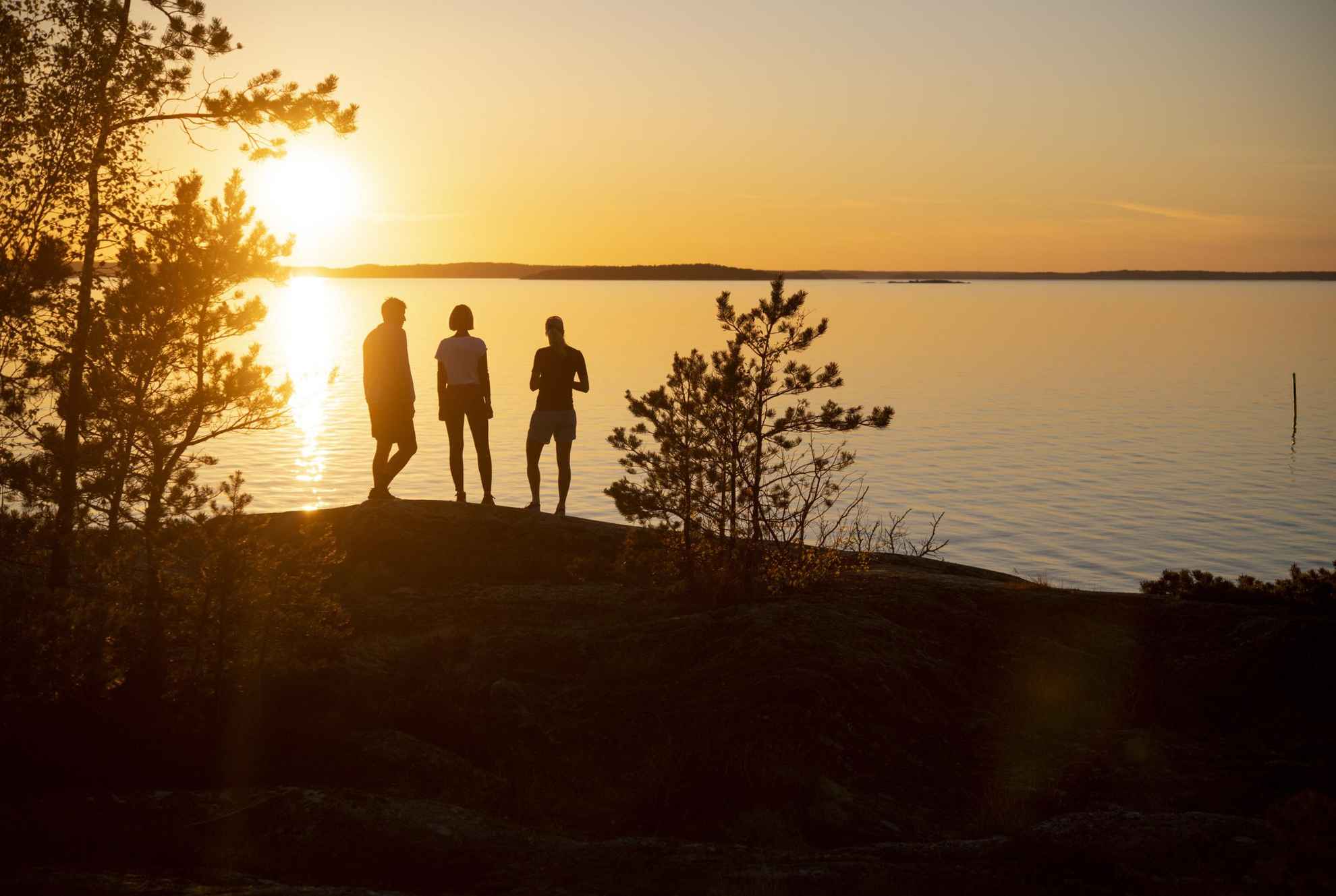 Drei Personen stehen am Meer und schauen sich den Sonnenuntergang auf einer Insel im Stockholmer Schärengarten an.