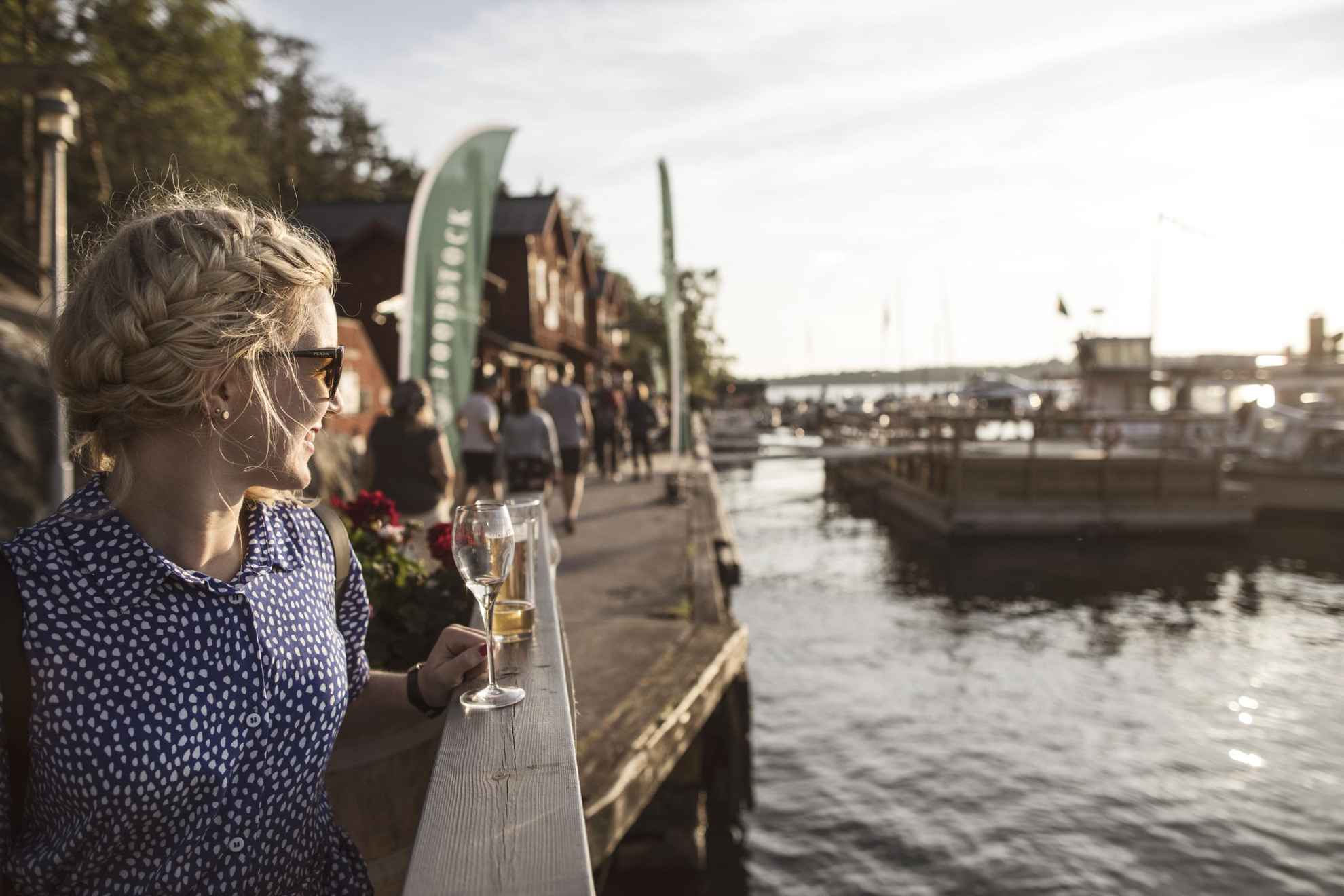 Eine Frau genießt einen Drink in der Sonne auf einem Steg am Meer. Unscharf im Hintergrund der Jachthafen und die Leute, die dort herumlaufen.