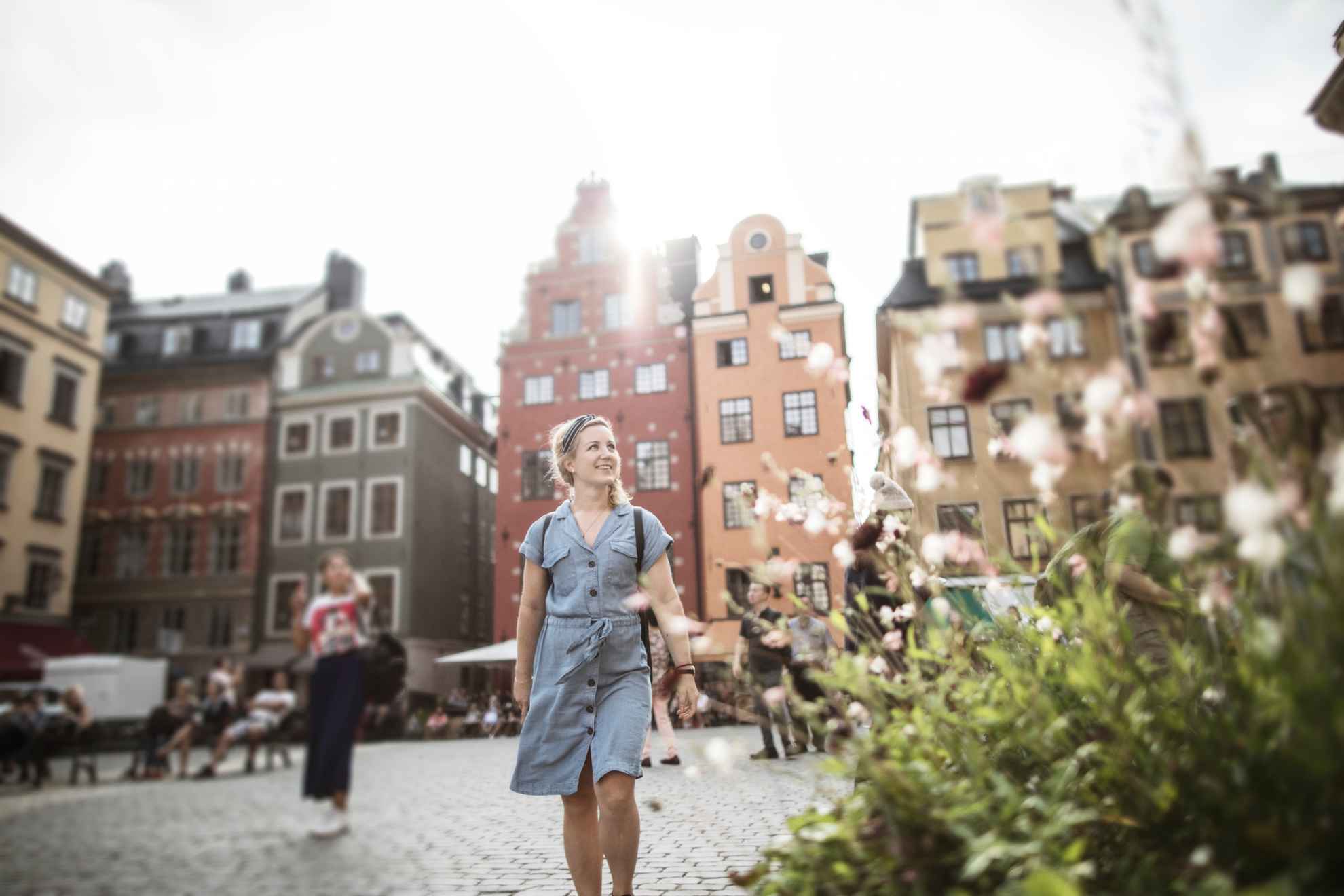 Eine Frau geht auf einem Platz in der Altstadt spazieren. Im Hintergrund siehst du alte Steinhäuser und im Vordergrund steht eine blühende Pflanze.