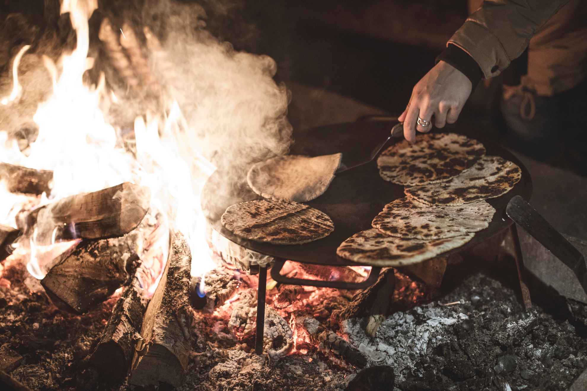Bake traditional Sámi bread, gahkku, on a flat iron plate over an open fire. A hand is flipping the bread.