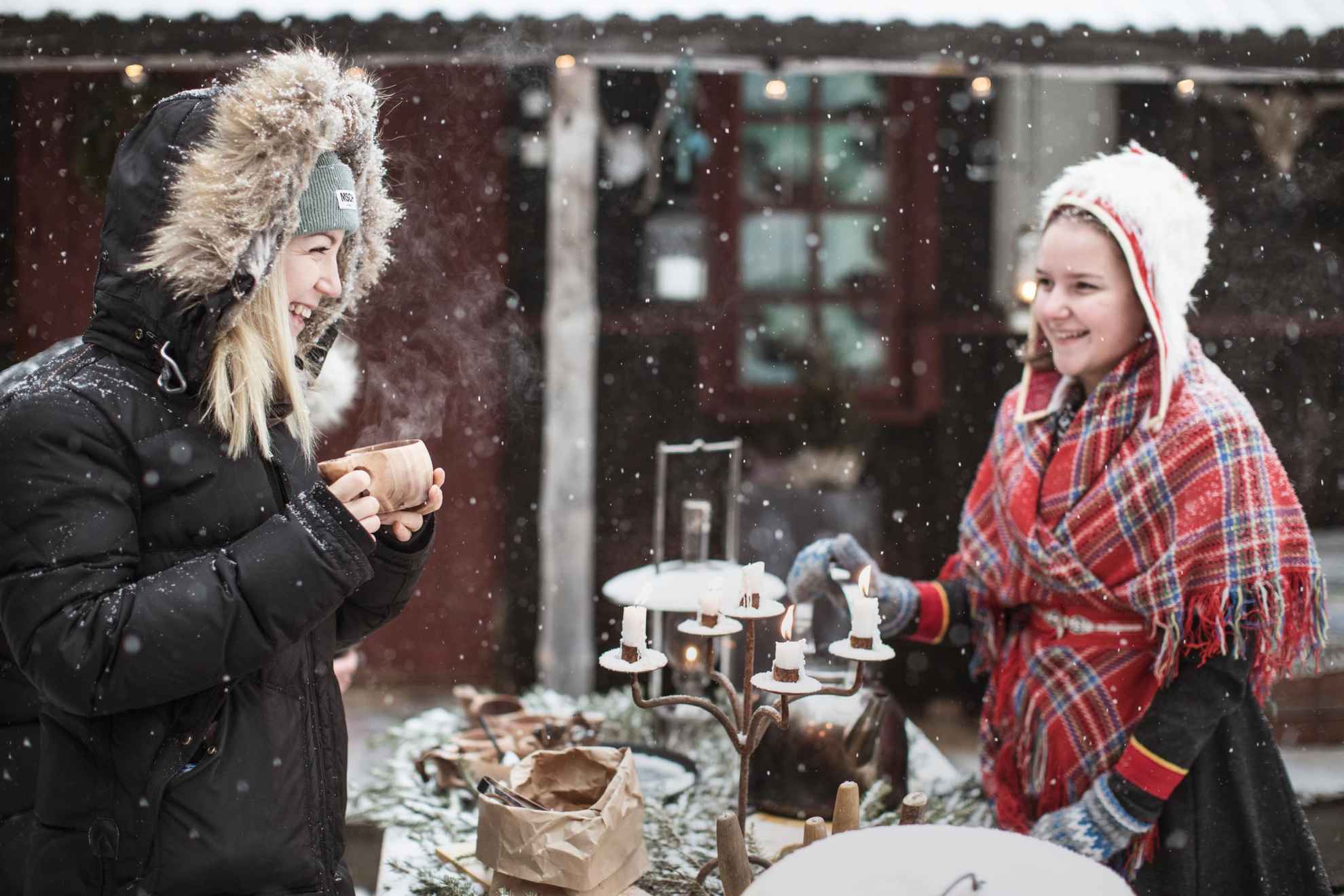 Zwei Frauen stehen an einem Tisch und halten eine Fika im Freien im Schnee. Auf dem Tisch steht ein alter Kandelaber mit brennenden Kerzen. Eine der beiden Frauen trägt eine samische Tracht. Es schneit.