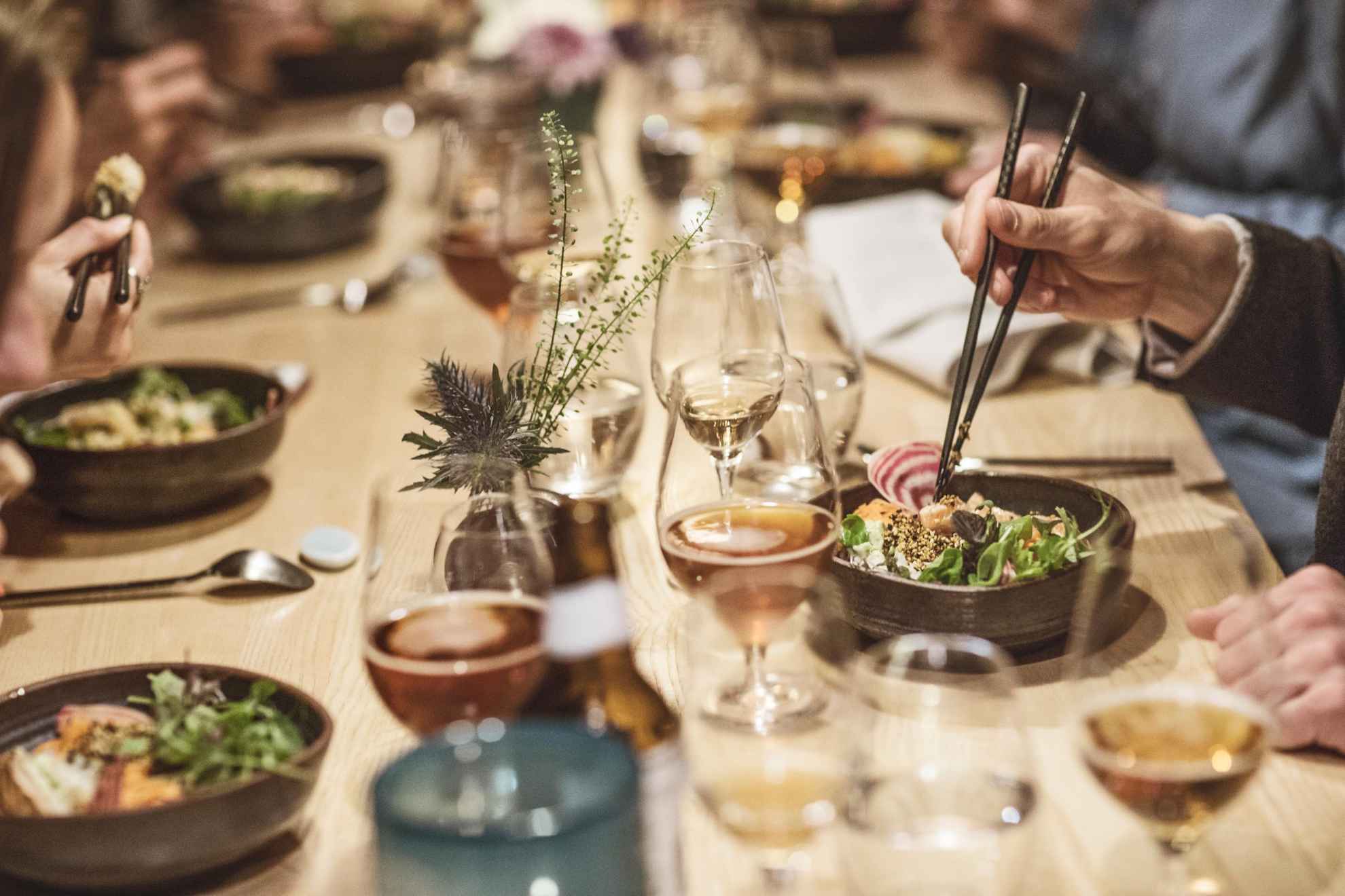 Ein Esstisch mit mehreren Tellern und Gläsern. Menschen sitzen um den Tisch herum. Jemandes Hand, die Essstäbchen hält, ist sichtbar.