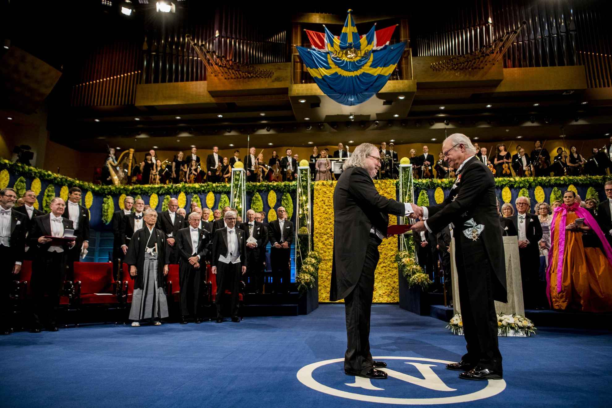 Ein männlicher Preisträger steht auf einer Bühne und nimmt seinen Preis vom schwedischen König Carl XVI. Gustaf entgegen.