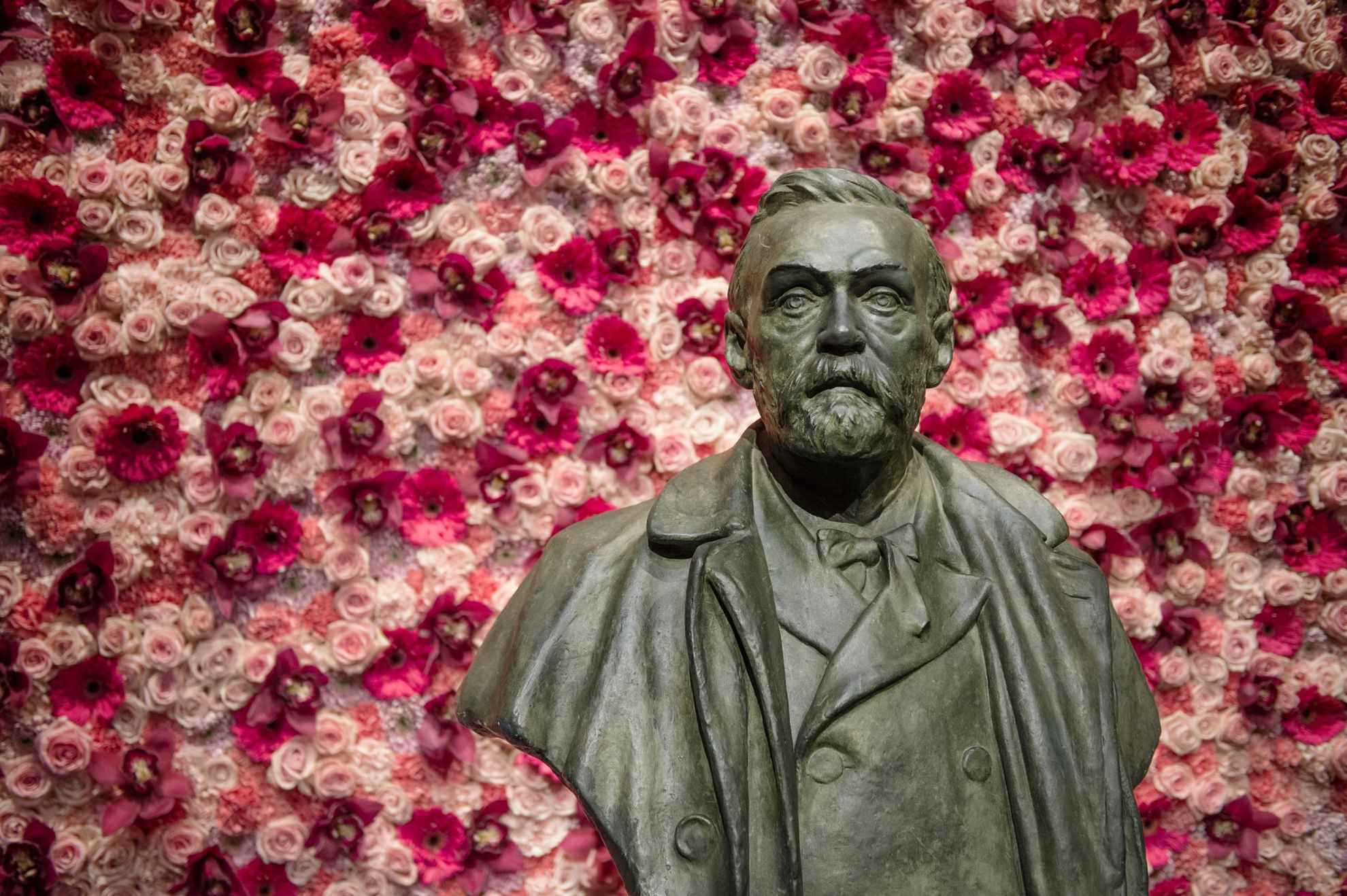 Eine Statue von Alfred Nobel vor einer mit rosa und weißen Blumen geschmückten Wand.