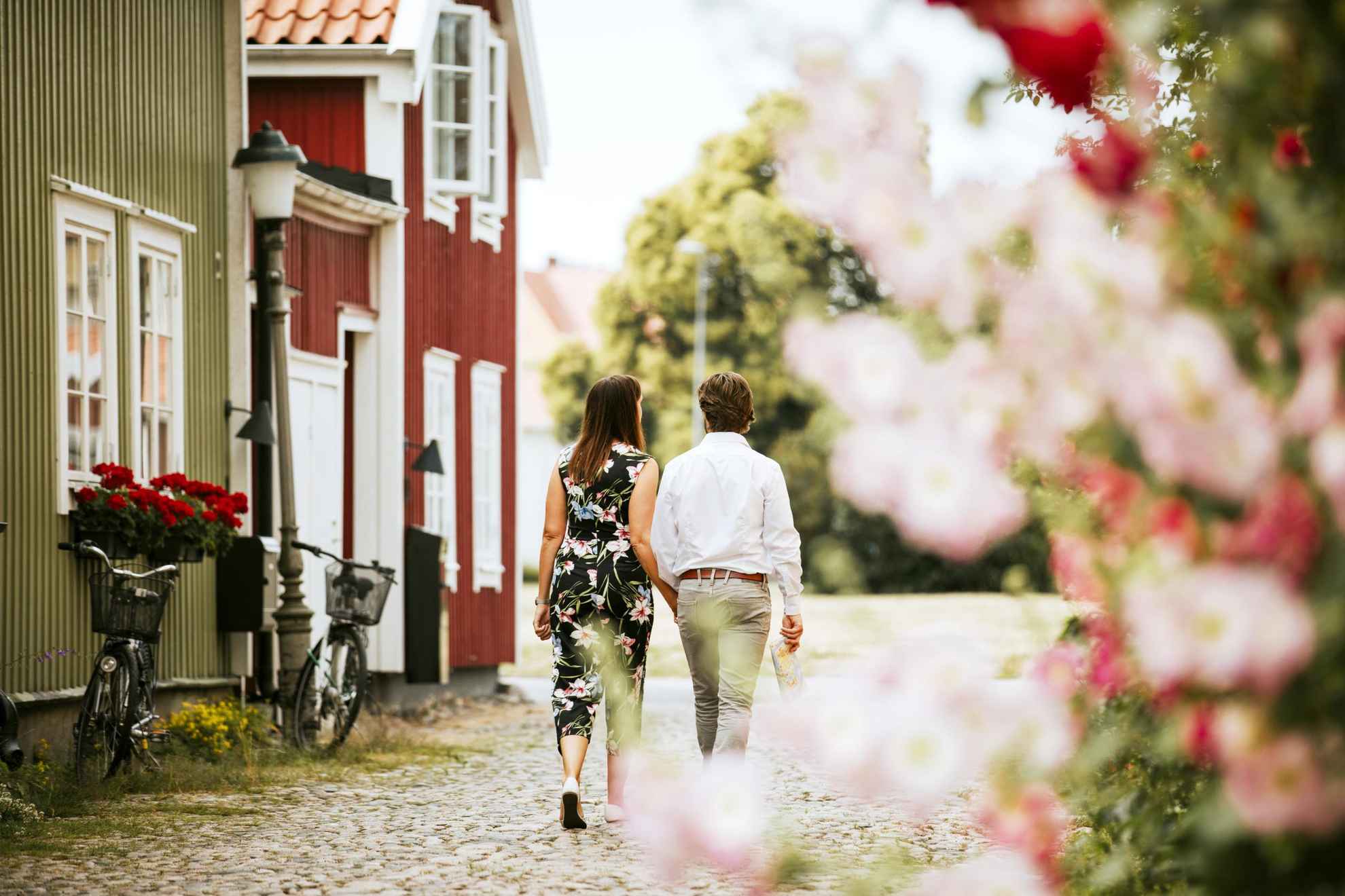 Der Rücken eines Paares, das neben einem roten und einem grünen Holzhaus in der Altstadt von Kalmar spazieren geht. Rosa Blumen unscharf im Vordergrund.