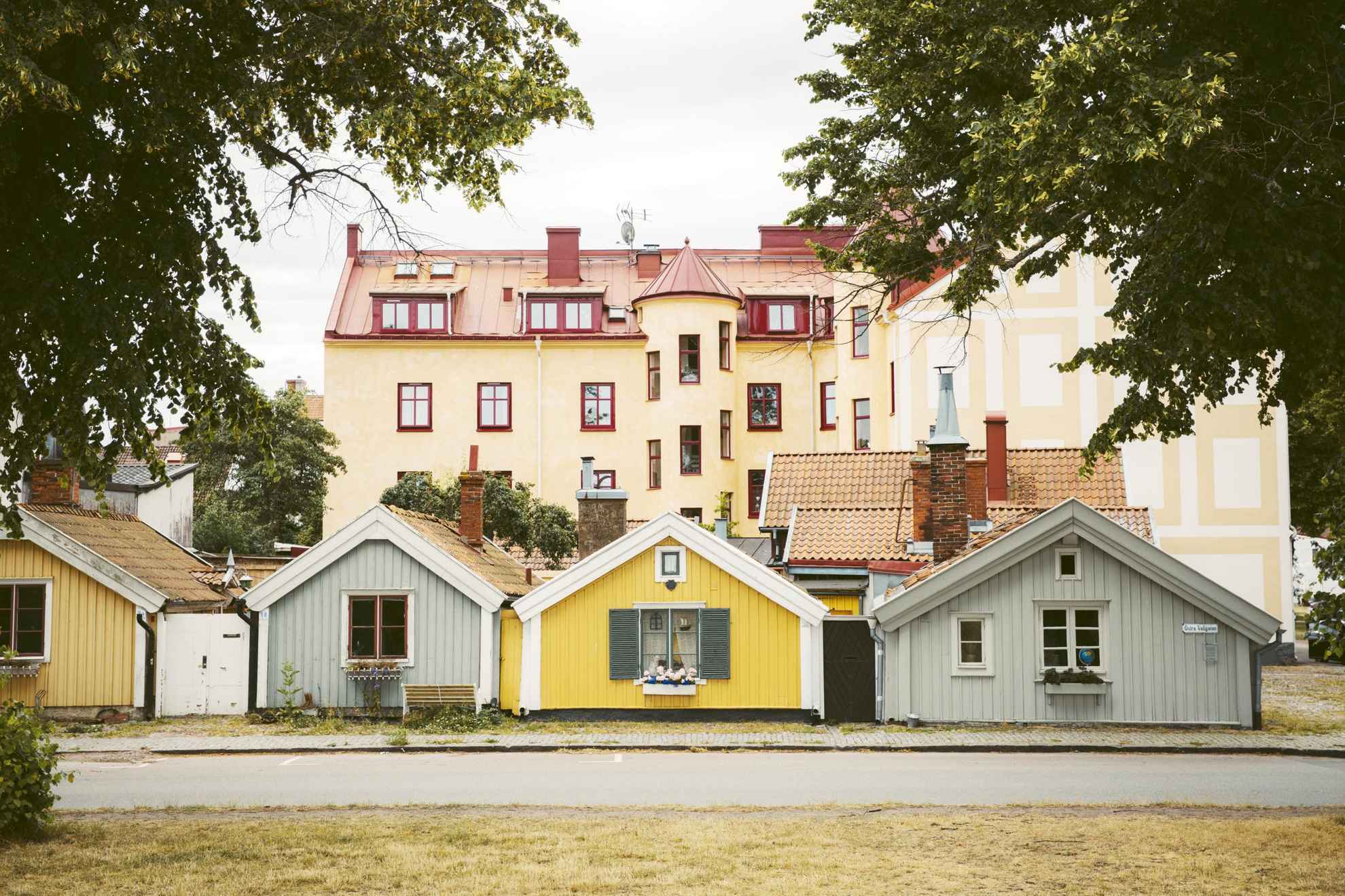 Vier kleine Holzhäuser in verschiedenen gelben und grauen Farben entlang einer Straße in der Altstadt. Dahinter ein großes hellgelbes Steinhaus.