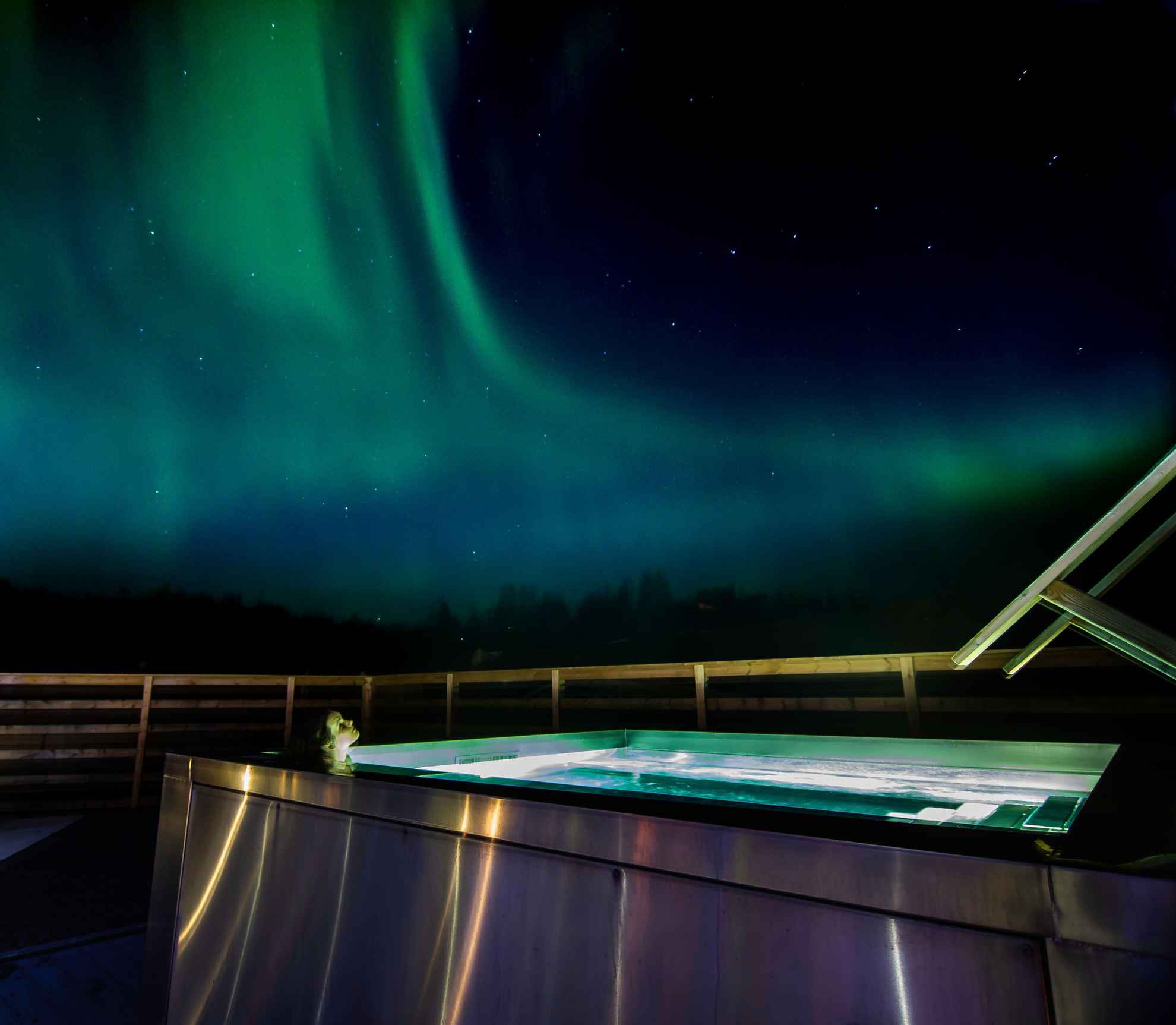 Eine Person sitzt in einem Whirlpool. Am Nachthimmel tanzen Nordlichter.