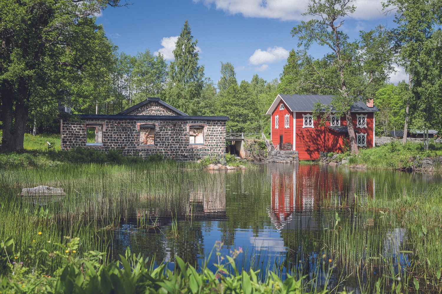 Ein rotes Holzhaus mit weißen Zierleisten und ein altes Steinhaus neben einem See im Sommer.