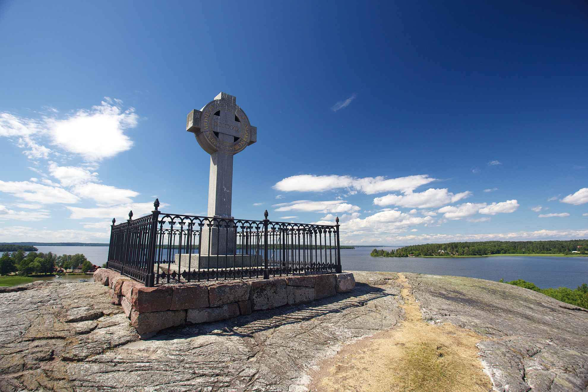 Das Ansgarkreuz steht auf einem kahlen Felsen. Im Hintergrund sind der See Mälaren, kleine Inseln und ein blauer Himmel zu sehen.