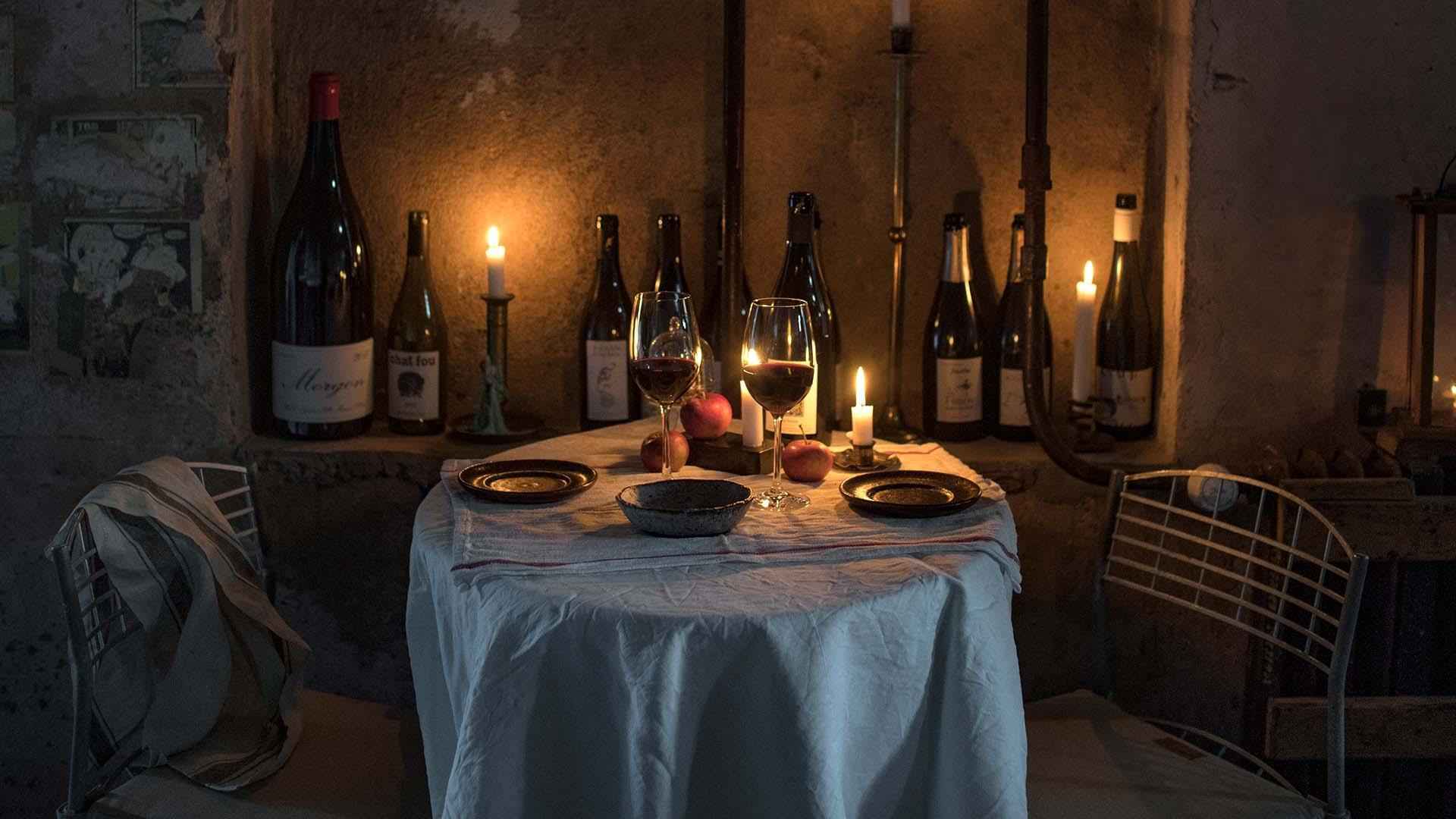Ein Tisch für zwei Personen mit einer weißen Tischdecke, Tellern und zwei Gläsern gefüllt mit Rotwein. Die Wand ist mit leeren Weinflaschen und brennenden Kerzen geschmückt.