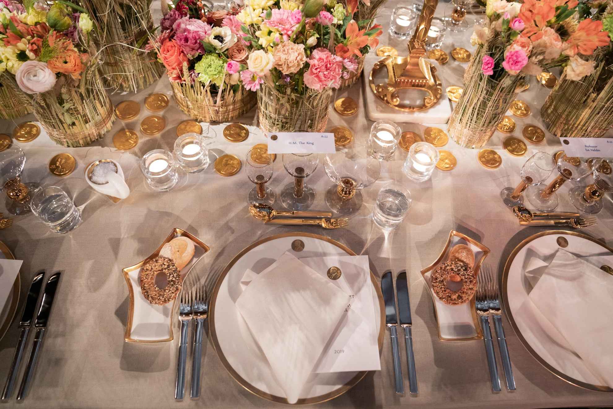 Ein Geschirr in Weiß mit goldenen Details wird auf einem Tisch mit einer weißen Tischdecke gedeckt. In der Mitte des Tisches stehen bunte Blumen in Vasen.