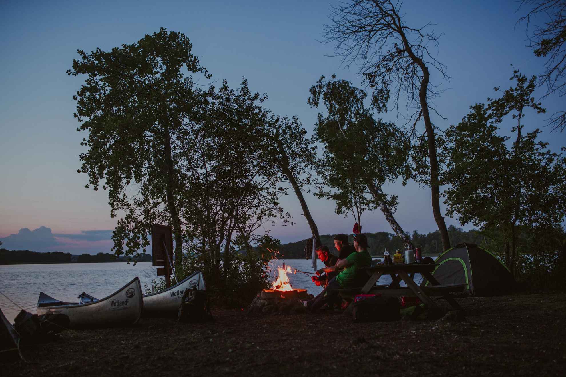 Menschen sitzen um ein Lagerfeuer verteilt am Ufer des Ivösjön es ist bereits dunkel. Am Ufer liegen zwei Kanus.