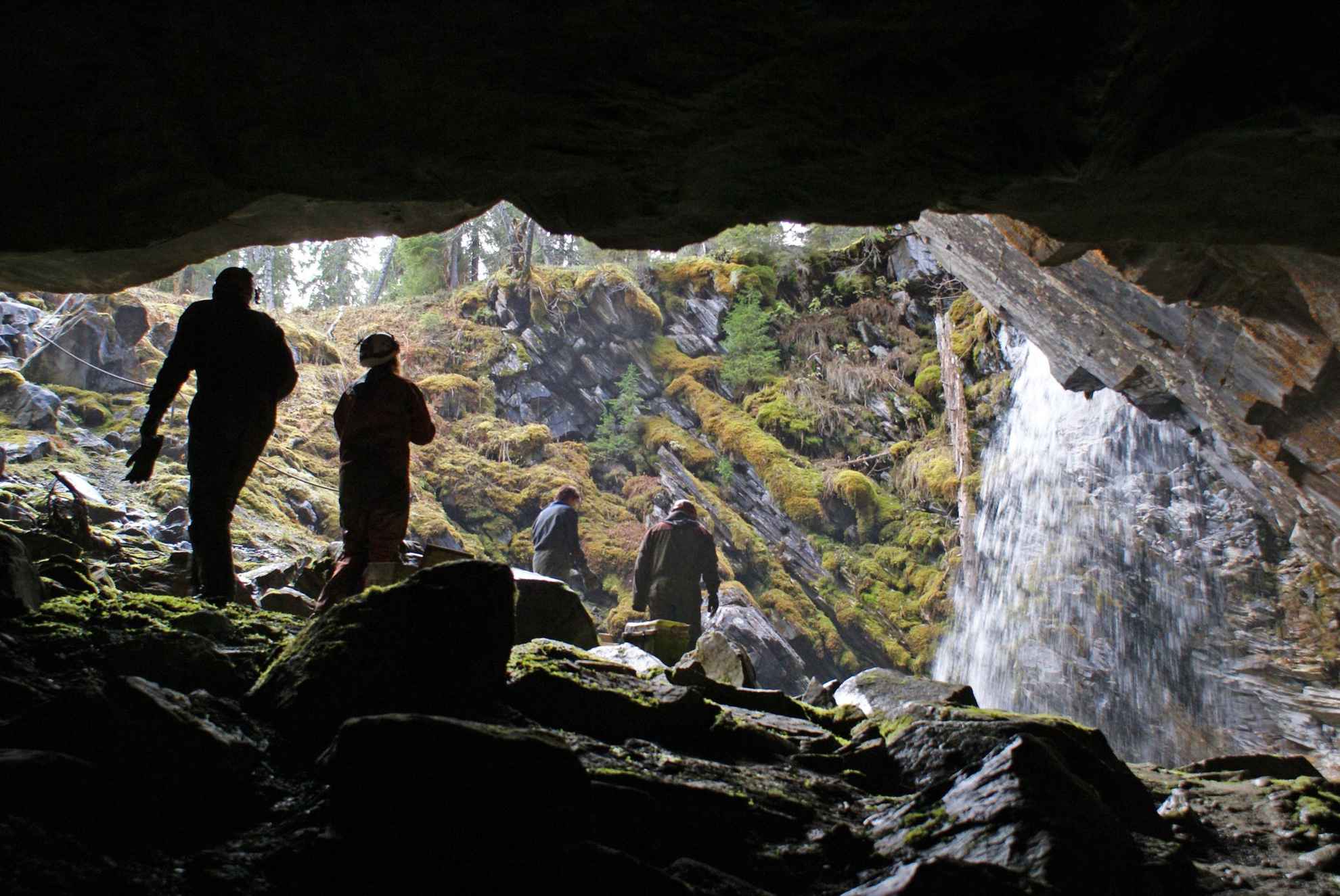 Ein Blick aus einer Höhle auf eine Lichtung mit vier Personen, die auf einen Wasserfall zugehen.