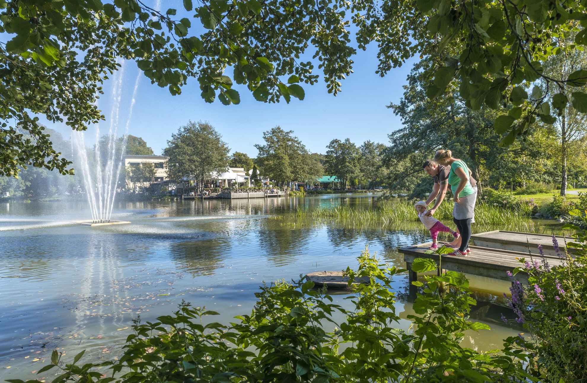 Ein Paar mit einem kleinen Kind steht auf einem Steg neben einem Teich mit Springbrunnen, umgeben von Grünflächen im Park Slottsskogen. Im Hintergrund sieht man ein Außenrestaurant.