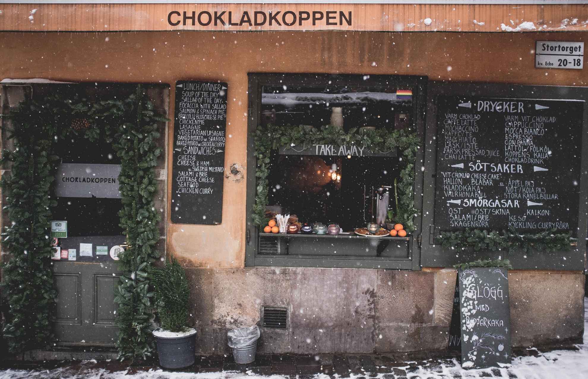 Aussenansicht des Café Chokladkoppen in Stockholm an einem Wintertag. Es schneit draußen und das Fenster des Cafes ist mit Tannenzweigen verziert.