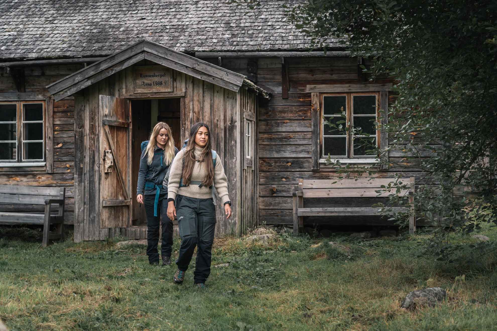 Zwei wandernde Frauen verlassen eine Holzhütte im Wald.