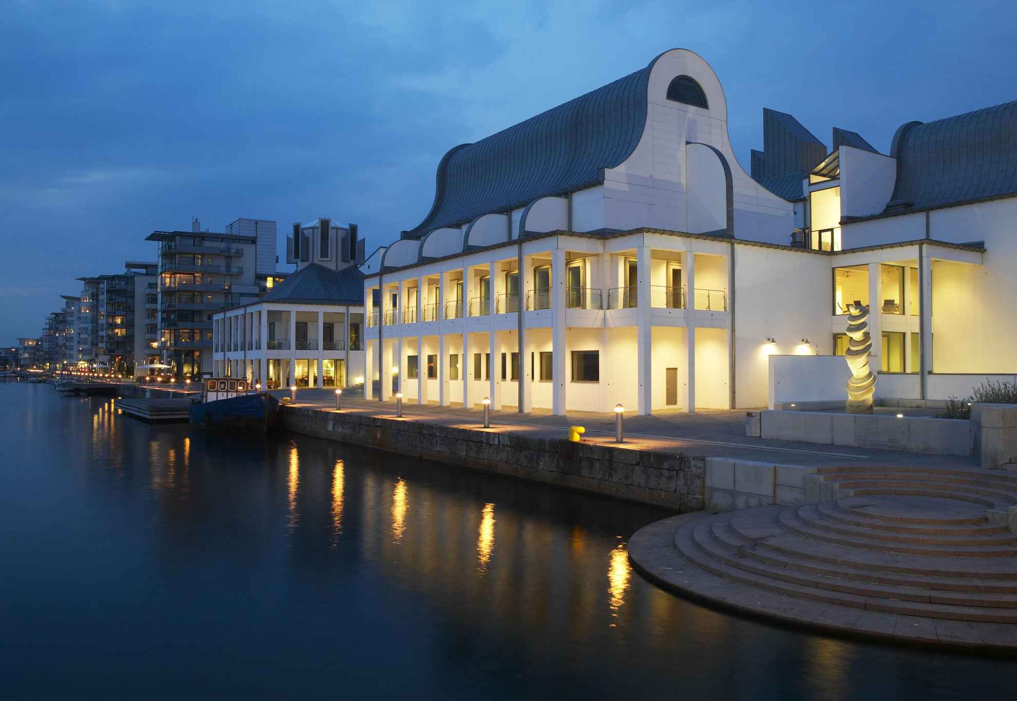 Dunkers Kulturzentrum im Abendlicht und mit Lampen an der Vorderseite, direkt am Meer an einem Jachthafen gelegen. Im Hintergrund siehst du Gebäude und Boote am Ufer.