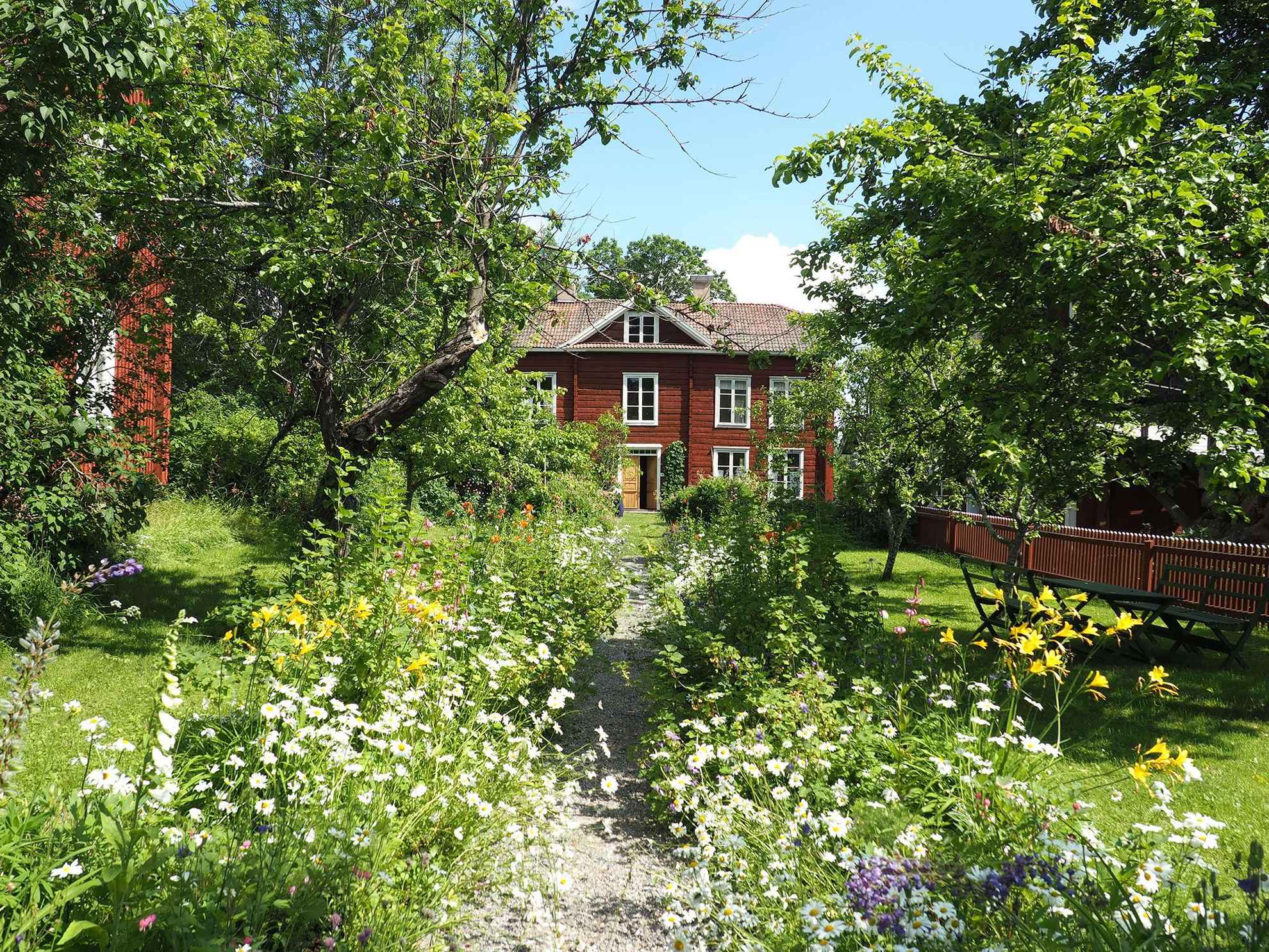 Eine Schotterstraße mit blühenden Blumen auf jeder Seite führt zu einem roten Holzhaus. Auf dem grünen Rasen stehen Gartenmöbel und Bäume.