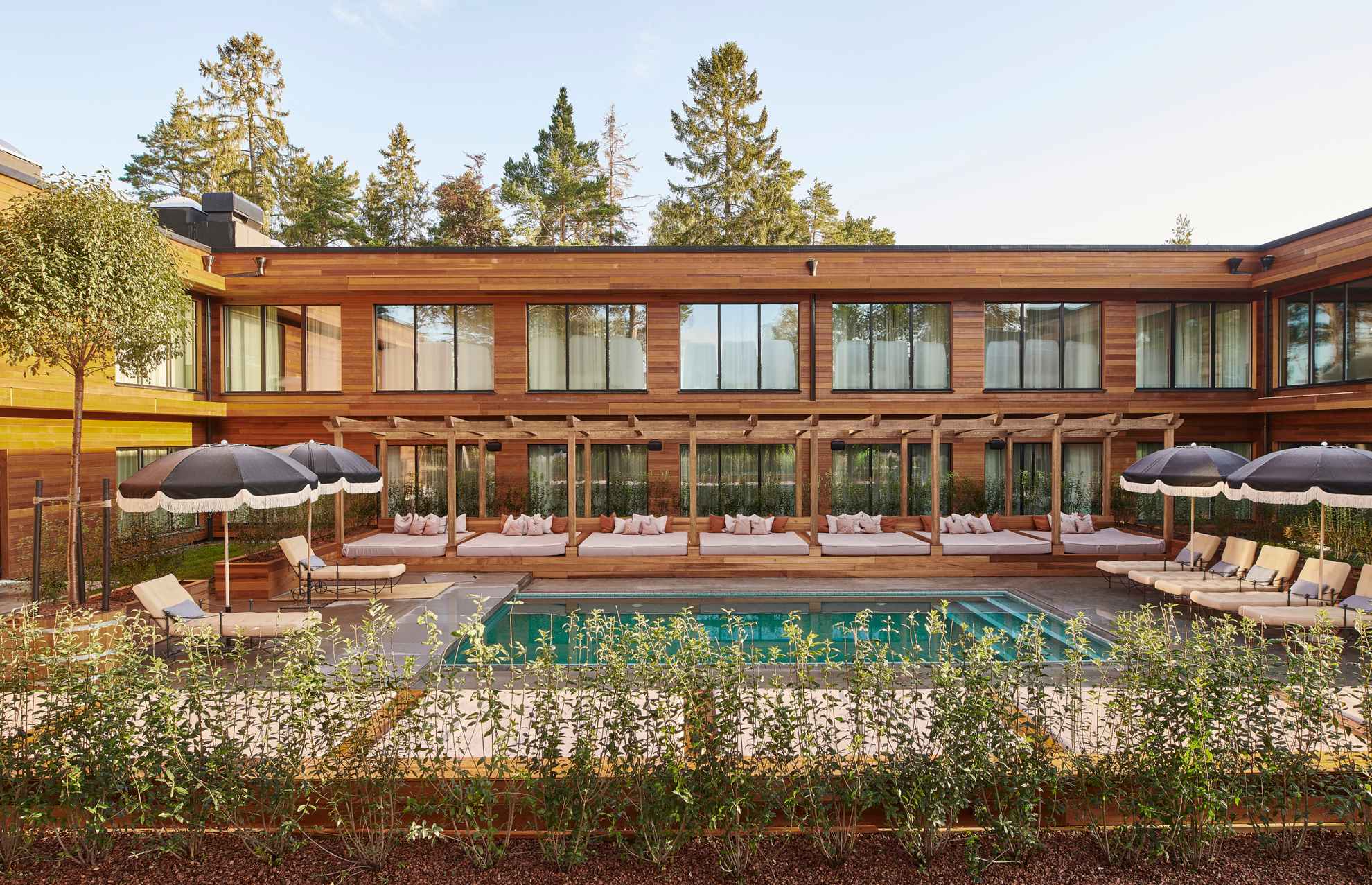 Ein Poolbereich mit Liegestühlen und Sonnenschirmen vor einem Holzhaus mit großen Fenstern.