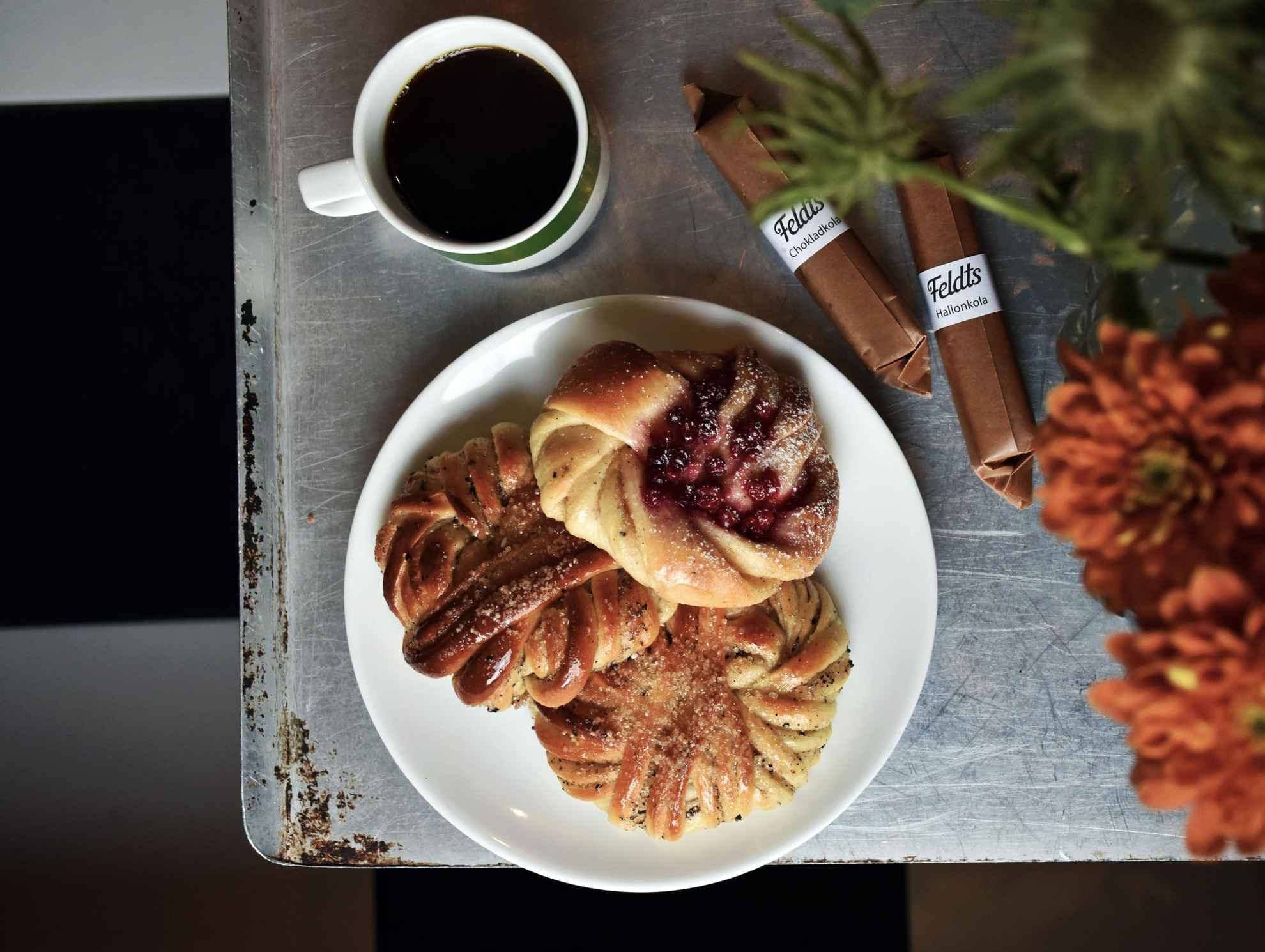 Close-up on a grey, wooden table with a cup of black coffee and three cardamom buns on a plate. Two packages of chocolate caramel is lying on the right side. Out of focus is a vase with orange flowers.