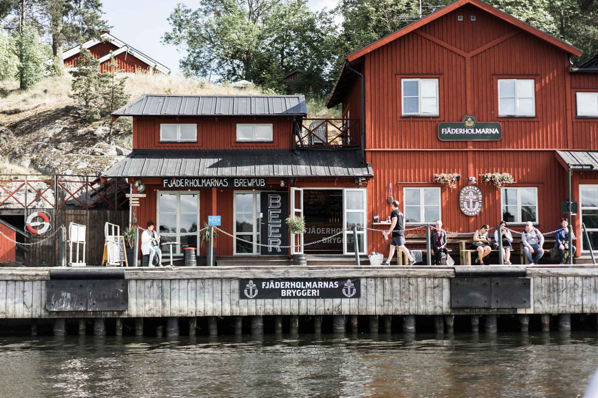 Fjäderholmarnas Brewery and Brewpub, located in a red wooden house at the sea side in the marina. People are sitting on benches outside.