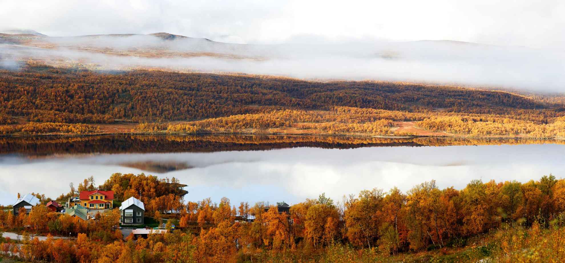 Eine schöne Herbstansicht eines Hotels an einem See in den Bergen. Die Bäume sind orange und auf der anderen Seite des Sees liegt Nebel über dem Berg.