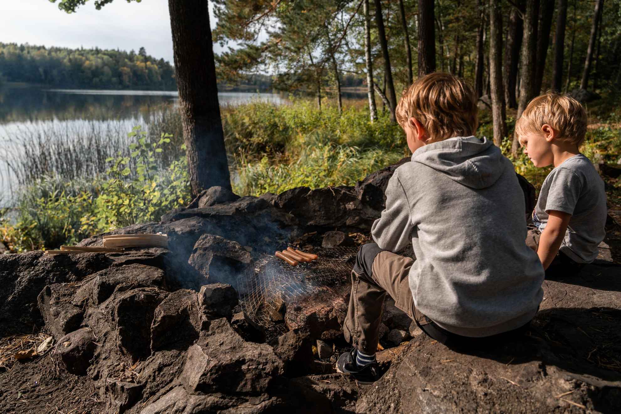 Zwei Kinder sitzen draußen in der Natur neben einem Feuer und grillen Würstchen.