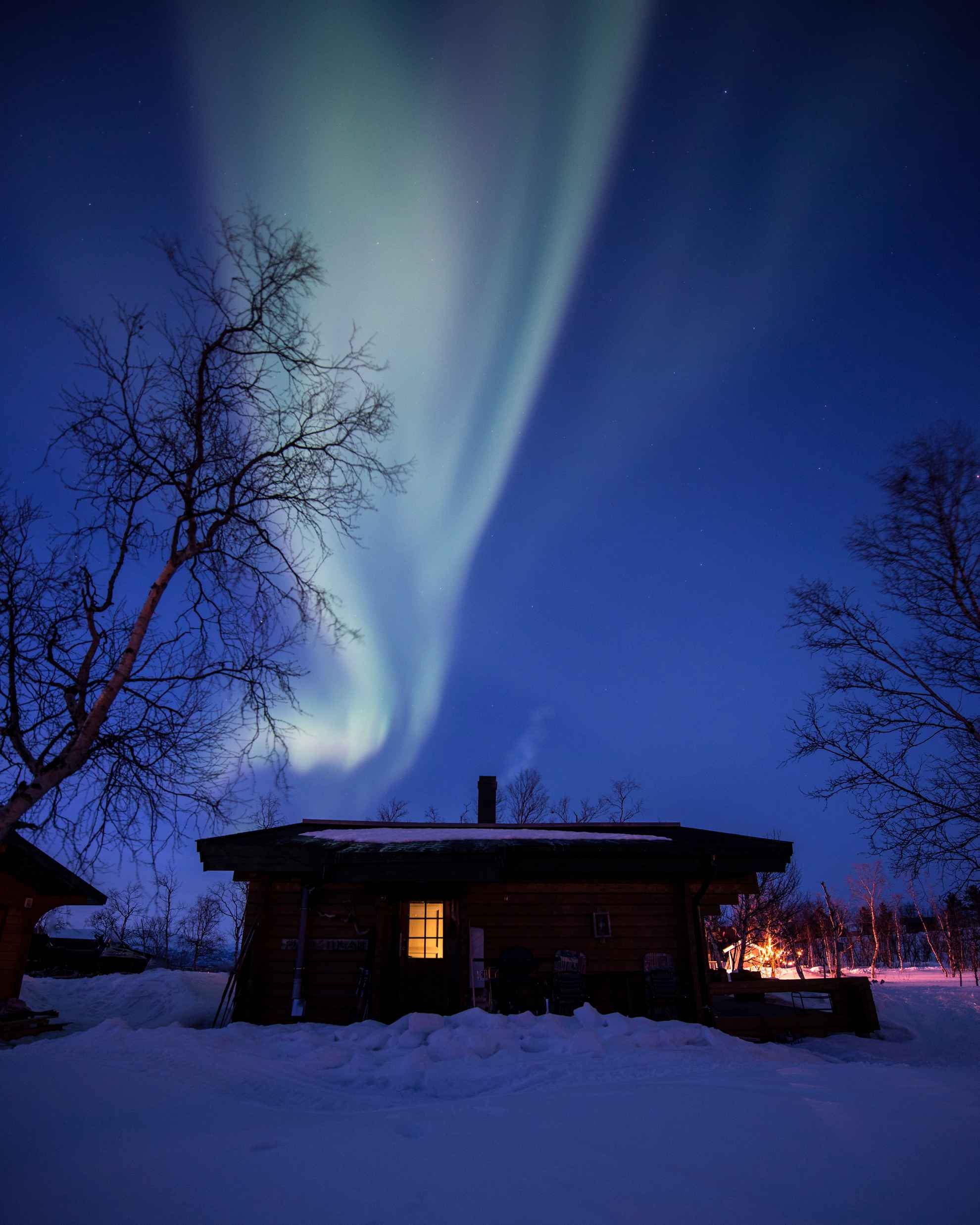 Das Nordlicht ist über einer Berghütte zu sehen. Es ist Winter und Schnee liegt auf dem Boden. Es ist Nacht und das Licht in der Hütte ist an.