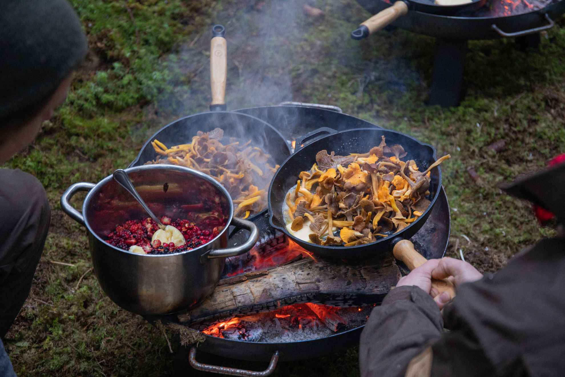 Menschen kochen in Guspfannen Pilzgerichte über dem offen Lagerfeuer im Wald.