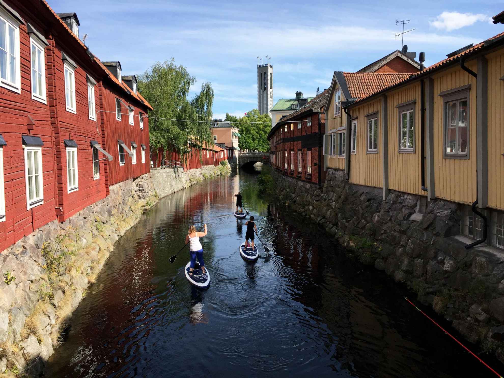 Drei Personen paddeln auf Stand-Up-Paddle-Boards in einem Kanal mit Häusern auf beiden Seiten.