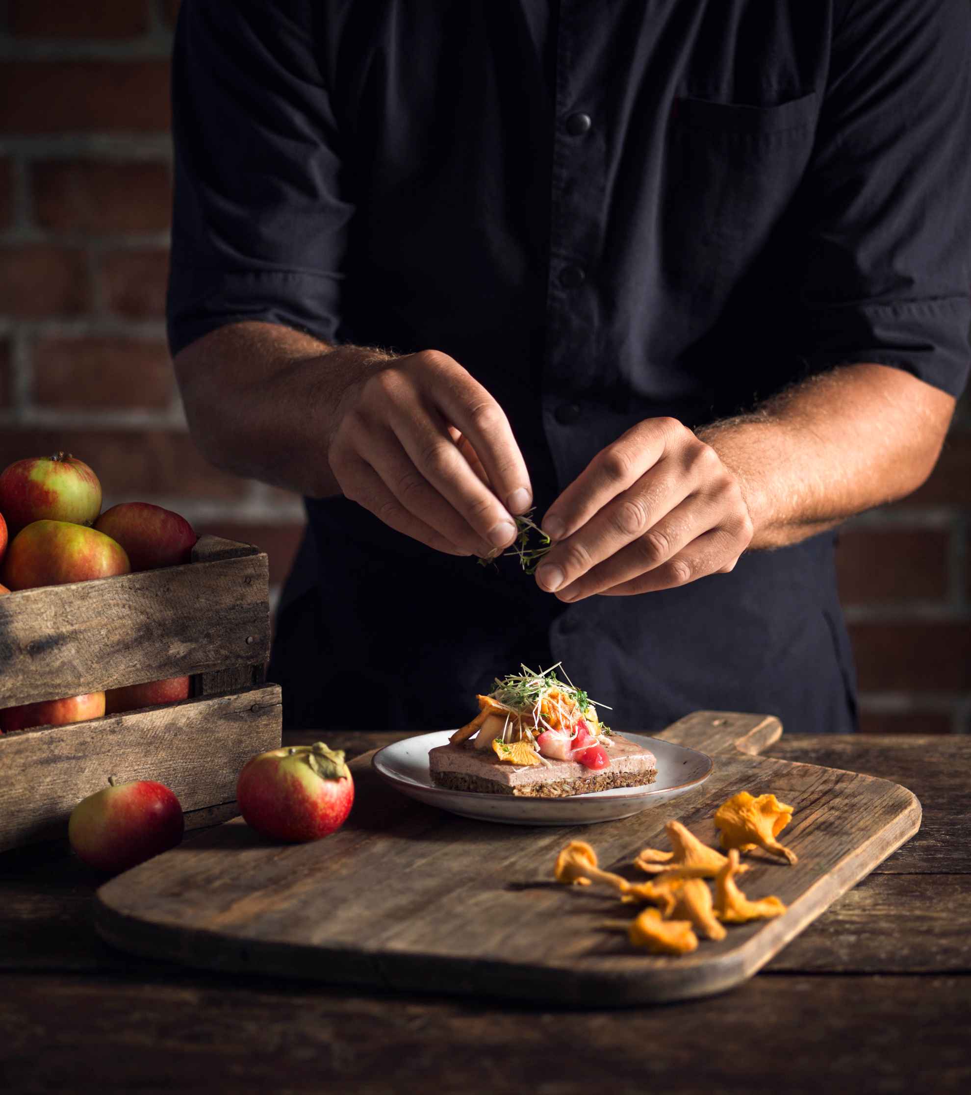The hands of a person is in focus, they are preparing a sandwich topped with chanterelles and vegetables. Apples and chanterelles lie on the wooden table