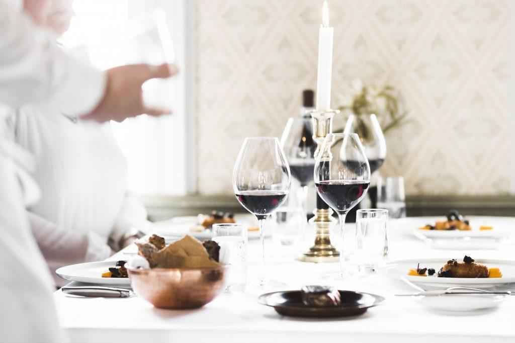 Ein mit einem weißen Tuch gedeckter Tisch, eine brennende Kerze, Teller mit Essen und Gläser mit Rotwein gefüllt. Im Vordergrund des Bildes steht ein Kellner.