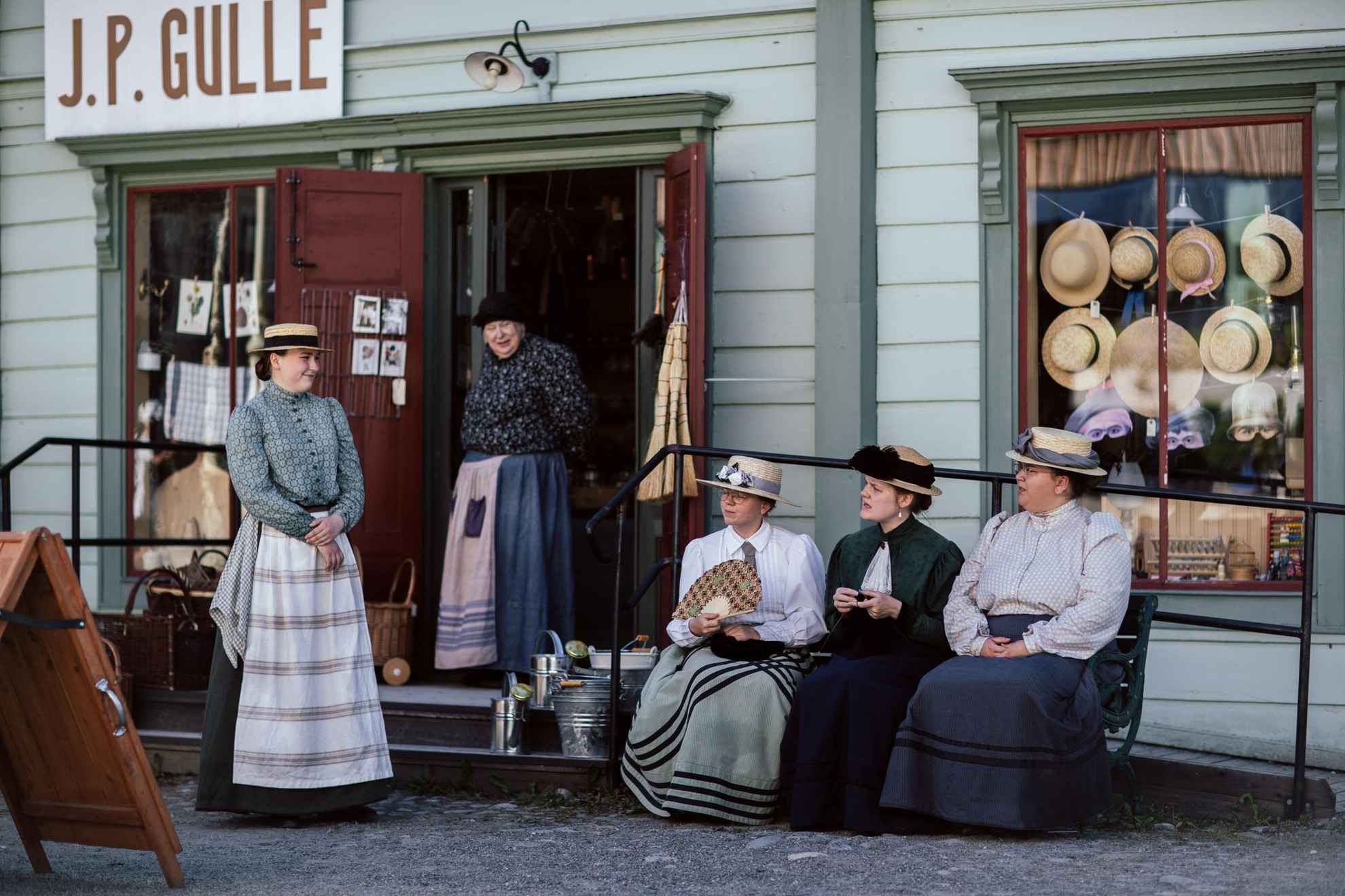 Frauen in Kleidern aus dem 19. Jahrhundert stehen und sitzen vor einem Haus in Jämtli.