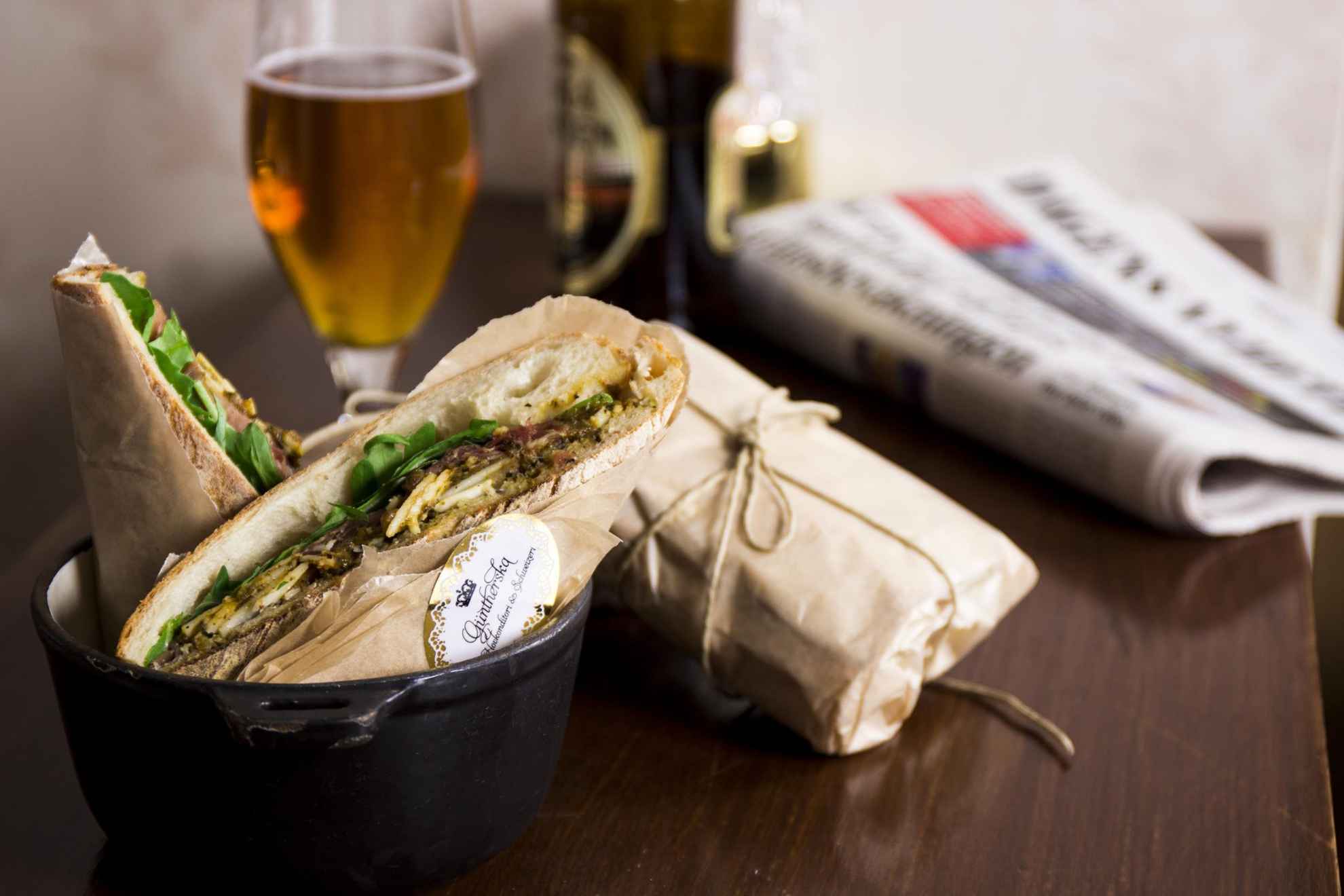 Nahaufnahme auf einem Tisch mit einem Sandwich, einem Bier und einer Zeitung.