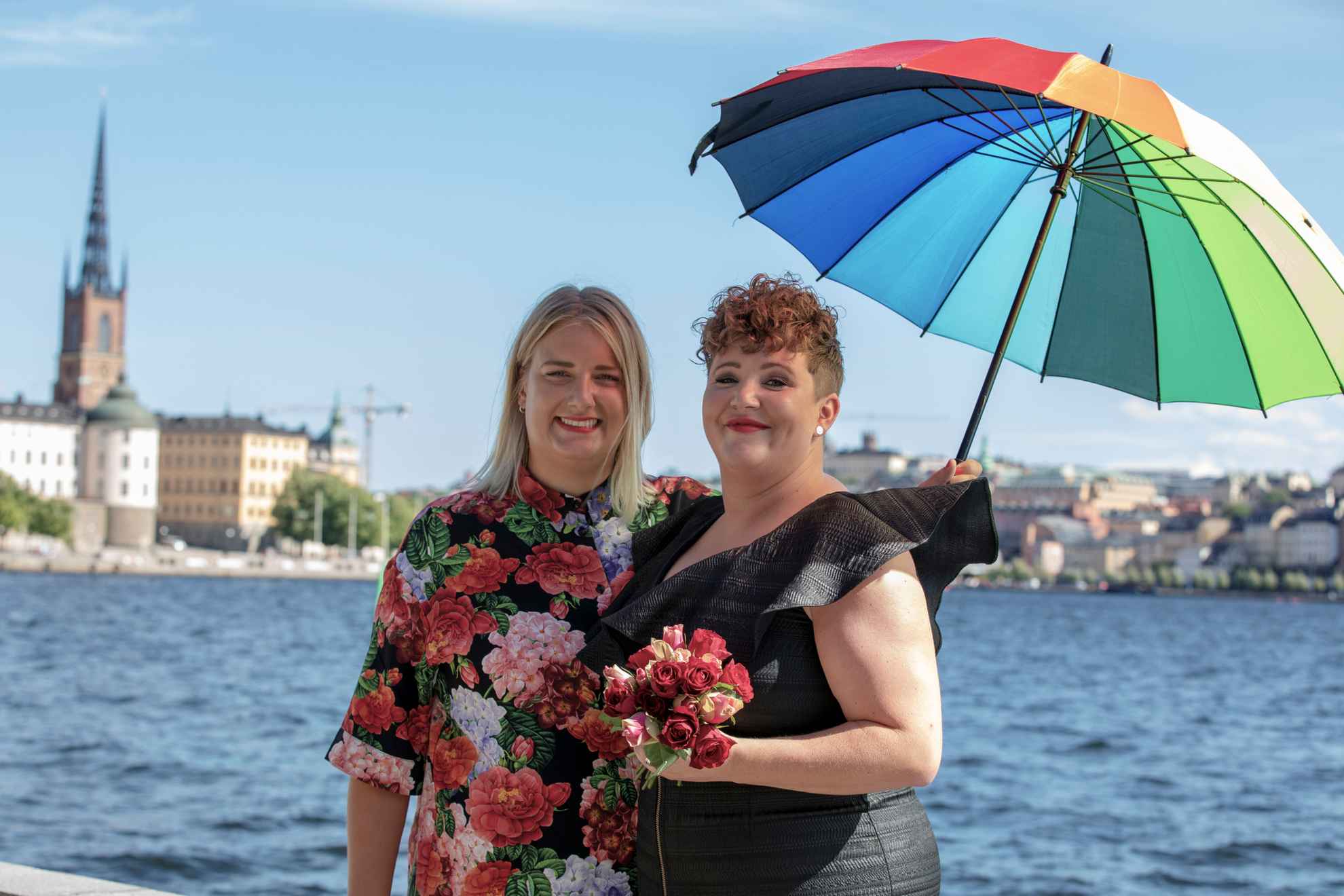 Zwei Frauen, eine in einem schwarzen Kleid und eine mit einem blumengemusterten Hemd, halten einen Regenschirm in den Farben des Regenbogens. Sie stehen am Wasser mit Blick auf die Stadt Stockholm im Hintergrund.