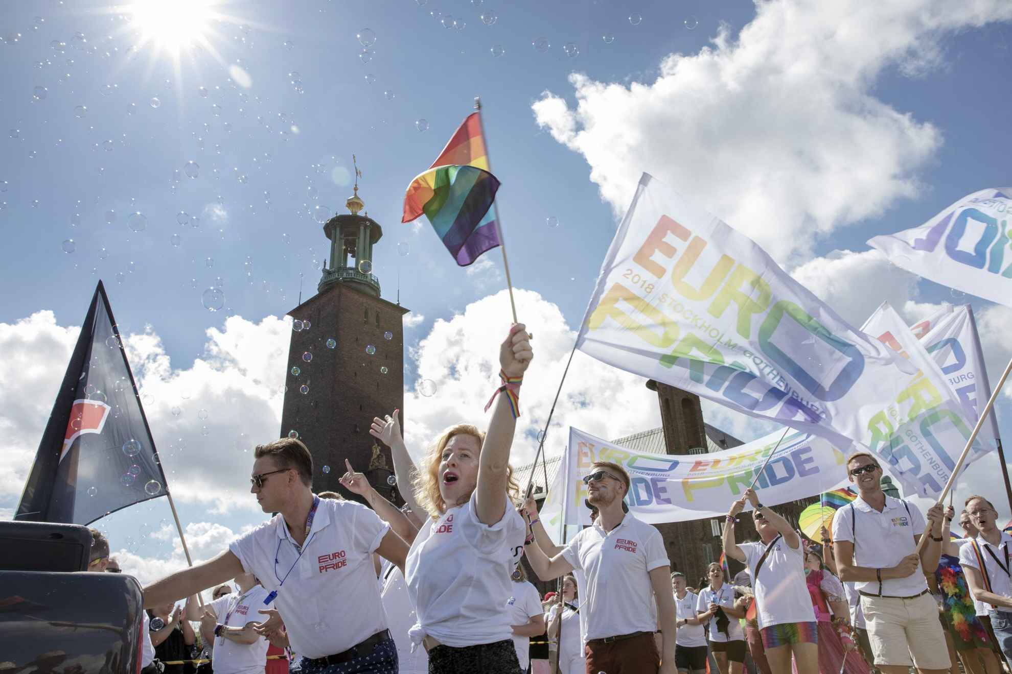 Mehrere Menschen gehen bei einer Pride-Parade. Sie haben Regenbogenfahnen und Fahnen mit dem Text Euro Pride. Im Hintergrund das Rathaus von Stockholm.