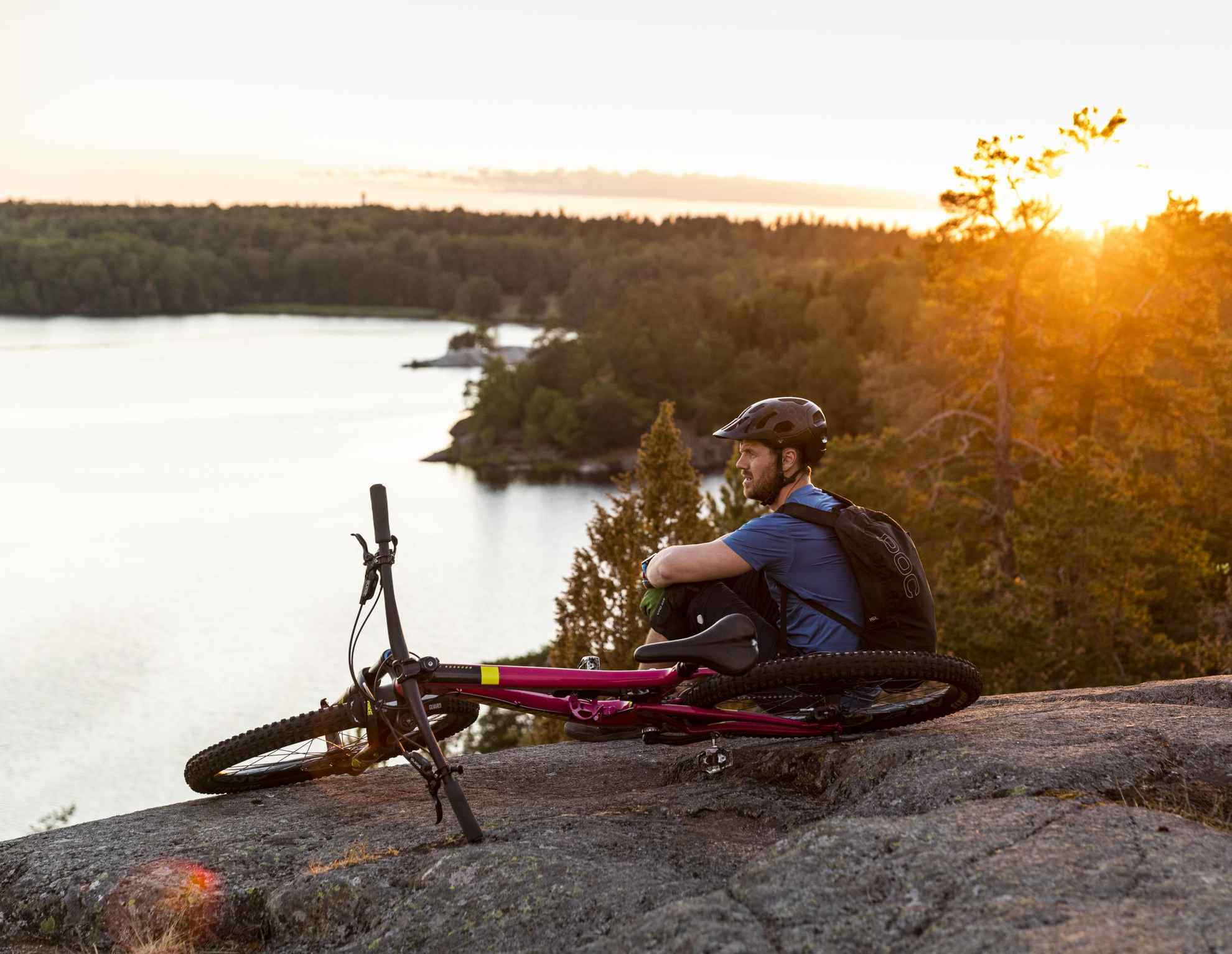 Ein Fahrradfahrer mit Helm sitzt auf einem Felsen, neben ihm sein Rad. Er blickt auf einen von Wald umgebenen See. Die Sonne geht unter.