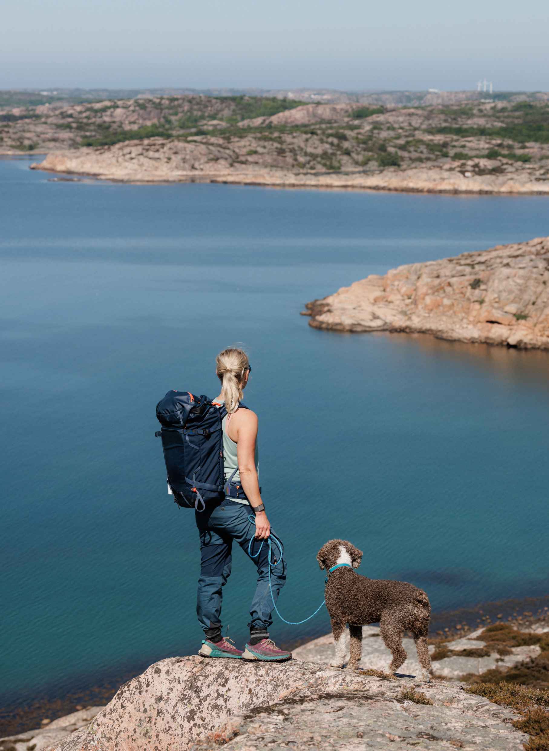 Eine Frau mit einem angeleinten Hund steht auf einer Klippe und blickt auf das Meer.