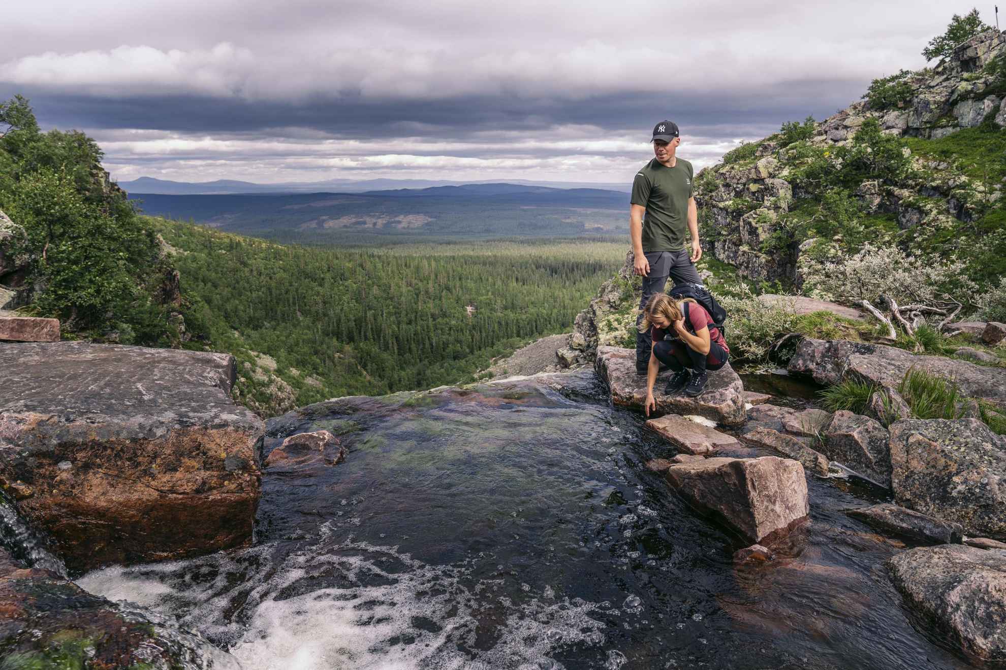 Eine Frau sitzt in der Hocke und ein Mann steht neben einer Quelle in den Bergen.Wälder und Berge befinden sich im Hintergrund.