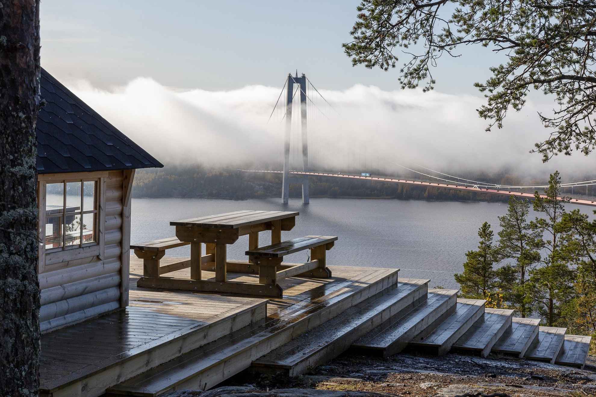 Ein hölzerner Picknicktisch mit Blick auf den Fluss Ångermanälven und die Hohe-Küsten-Brücke (Högakustenbron).