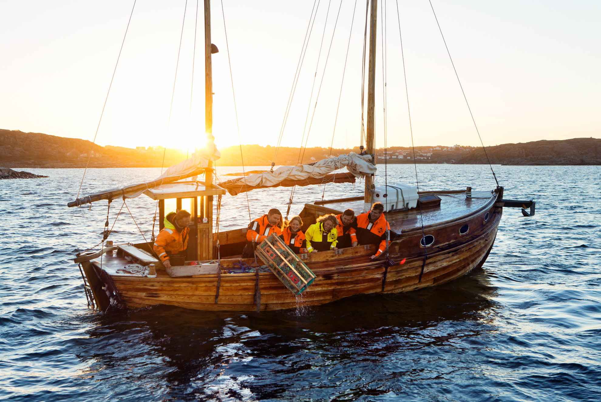 Ein hölzernes Segelboot mit sechs Personen, von denen eine gerade einen Hummerkäfig ins Meer wirft. Im Hintergrund geht die Sonne unter.