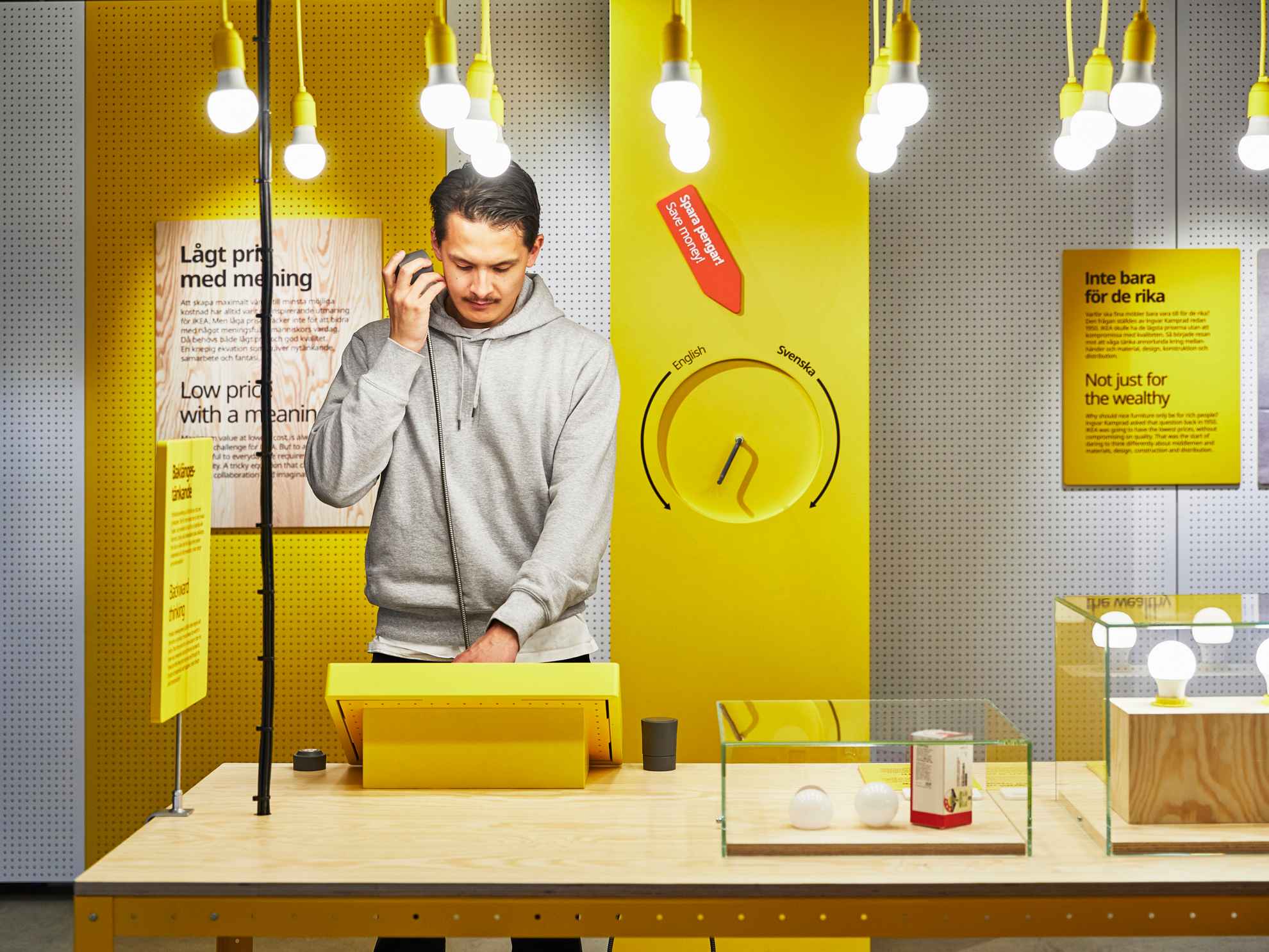 Ein Mann steht an einem Tisch und betrachtet eine gelbe Auslage in einer der Ausstellungen von Ikea. Von der Decke hängen Glühbirnen und auf dem Tisch stehen Glaskästen mit Glühbirnen darin.