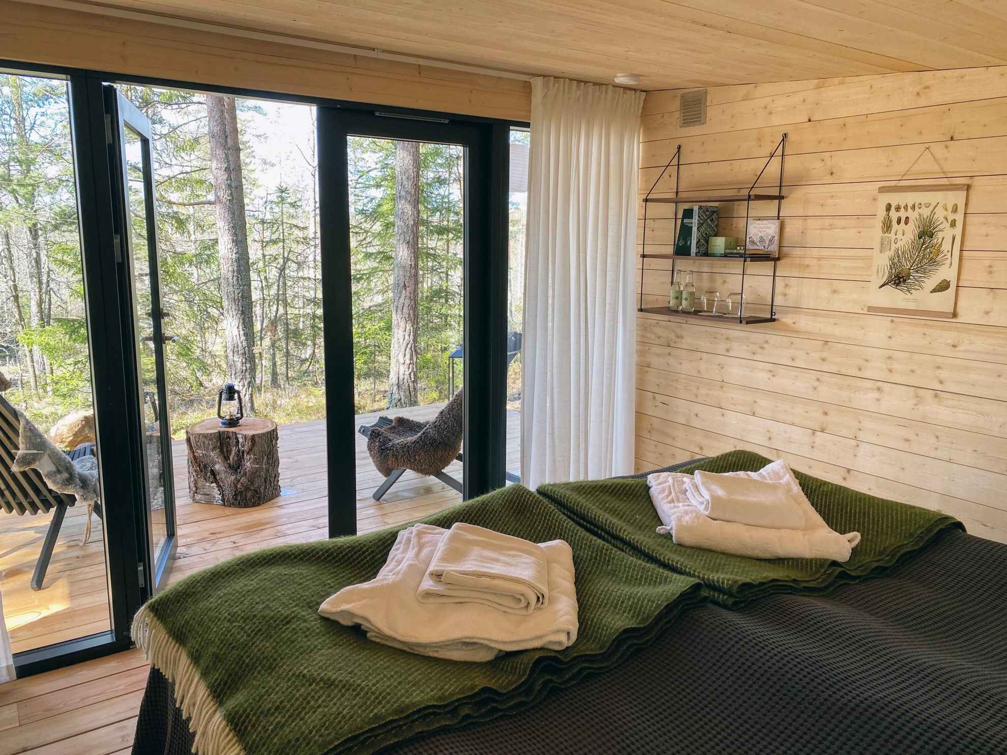 Zwei Betten befinden sich in einem Zimmer mit Holzwänden und -böden. Das Zimmer hat Glastüren zu einer Terrasse mit zwei Stühlen mit Blick in die Natur.
