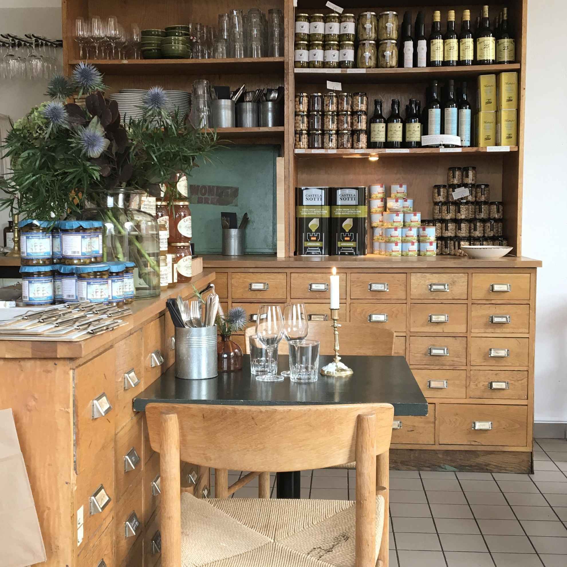 Ein mit Besteck gedeckter Holztisch des Restaurant Matateljén in Stockholm. Im Hintergund befindet sich ein Regal mit Lebensmitteln in Dosen und Gläsern.