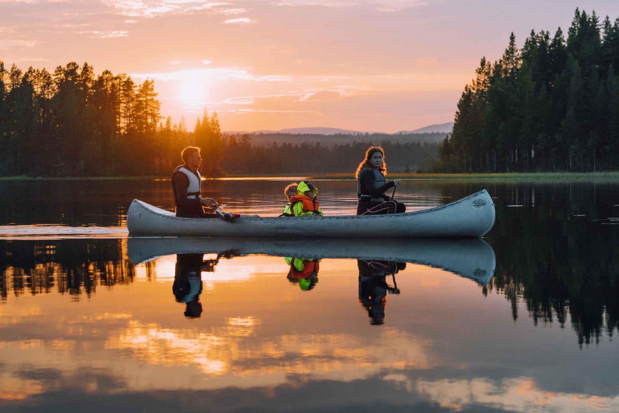 In einem Kanu sitzen 3 Personen, das Kanu gleitet entlang eines Sees im Sonnenuntergang